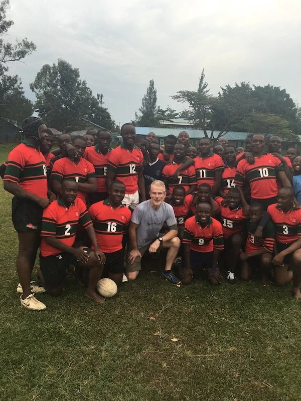 A group of young men in red and black rugby uniforms posing with a coach and a man in a gray shirt on a grassy field, with trees and buildings in the background.
