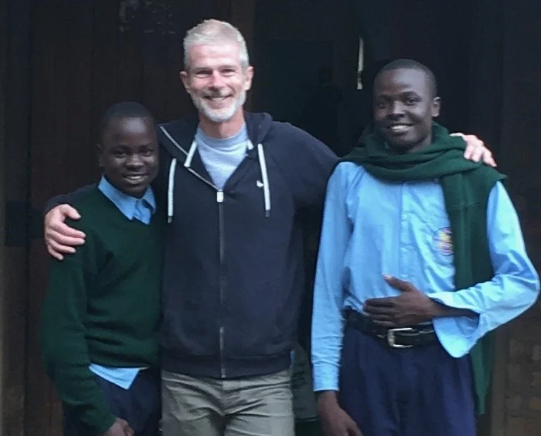 Three men smiling and standing together indoors, with the man in the center having his arms around the two men on either side.