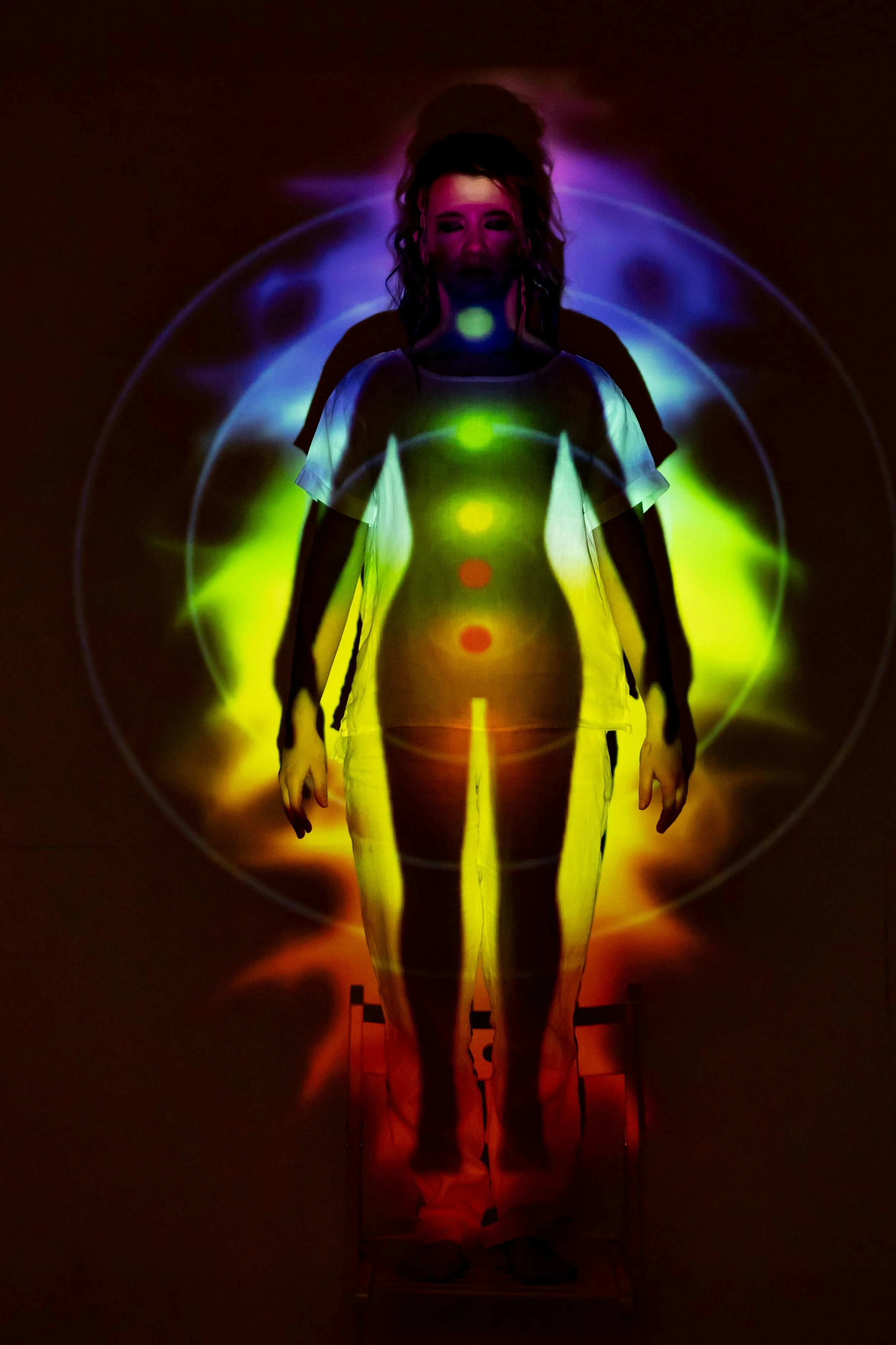 A woman standing on a chair with colorful light projections creating a visual effect on her and the background, resembling an abstract human outline.