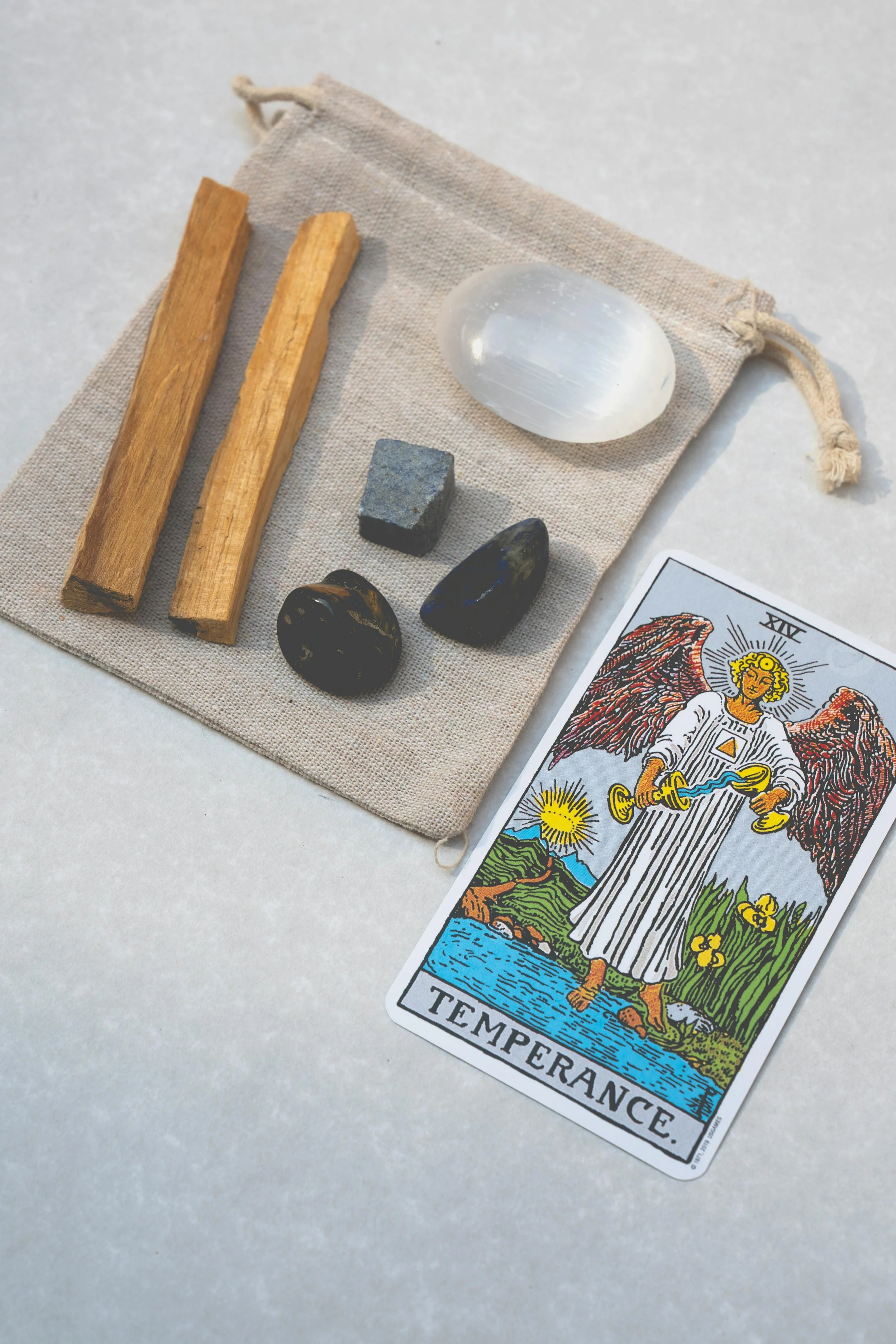 A tarot card labeled 'Temperance' placed beside a small cloth pouch containing two wooden sticks, a large moonstone, a smaller grey stone, and two dark polished stones on a light surface.