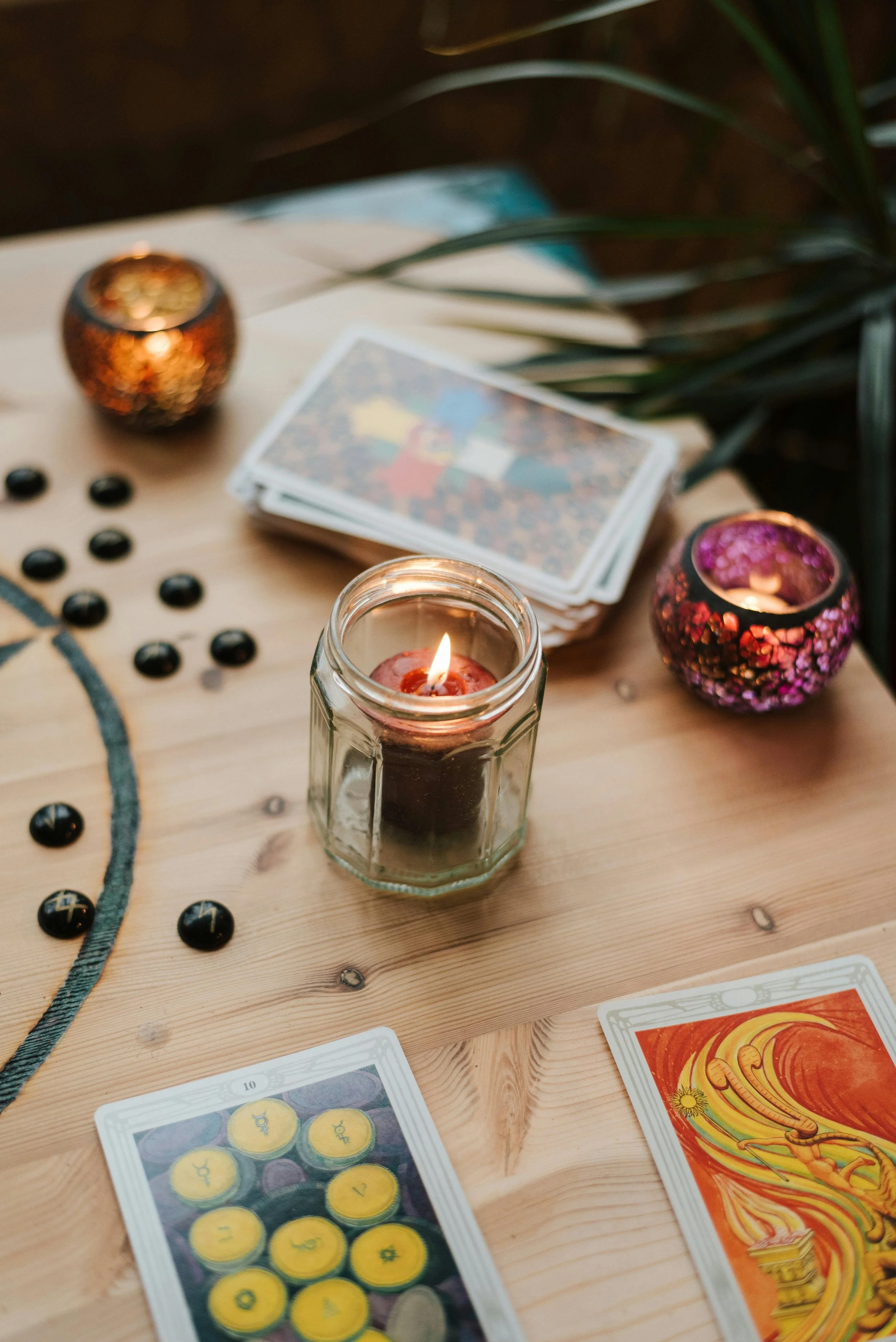 A wooden table with tarot cards, a lit candle in a glass jar, black game pieces, and small colorful candle holders, with green foliage nearby.