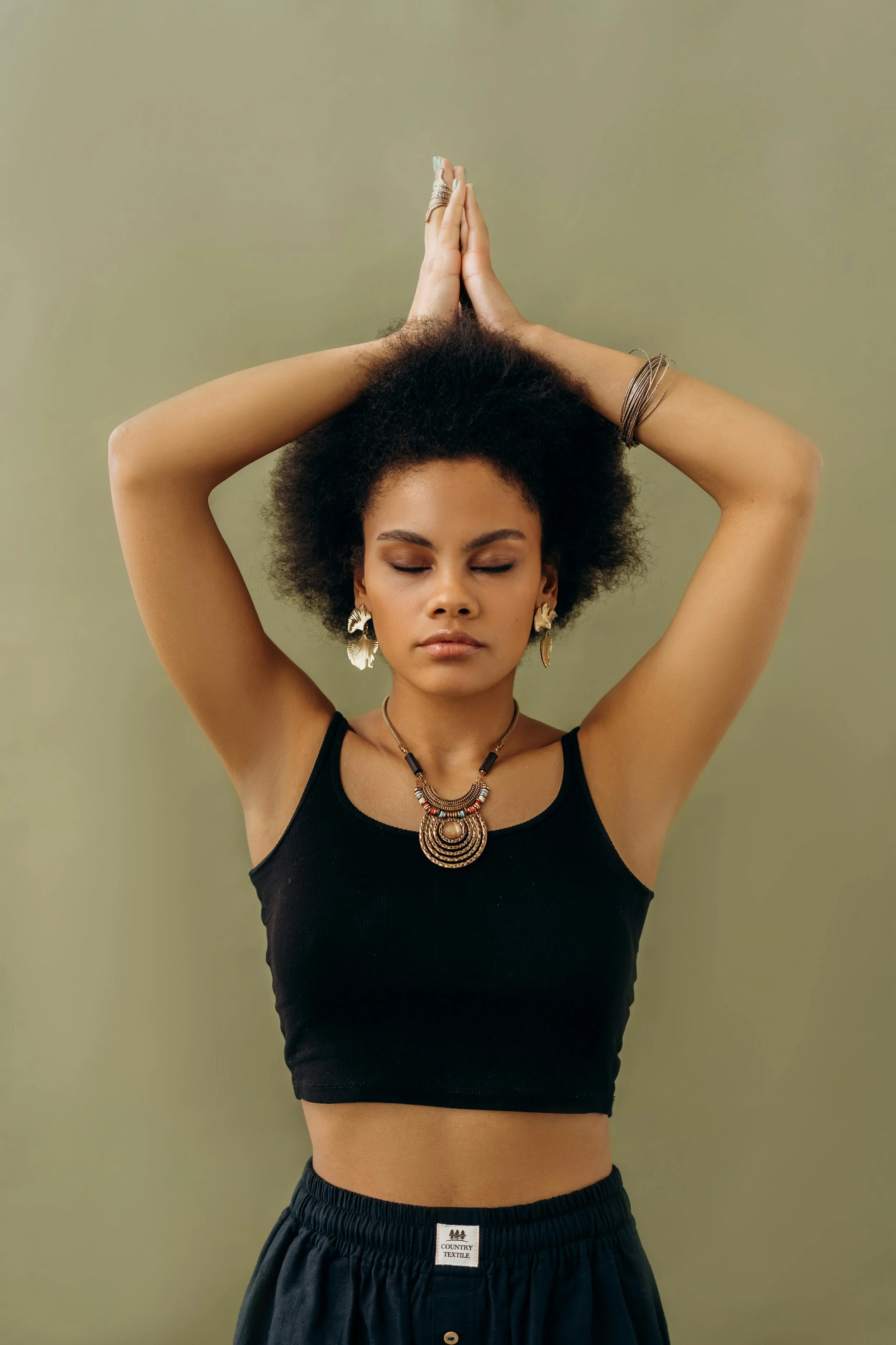 A woman with curly hair and medium skin tone posing with her eyes closed, wearing a black crop top, black pants, and jewelry including large earrings, a necklace, and bracelets, standing against a muted green background.