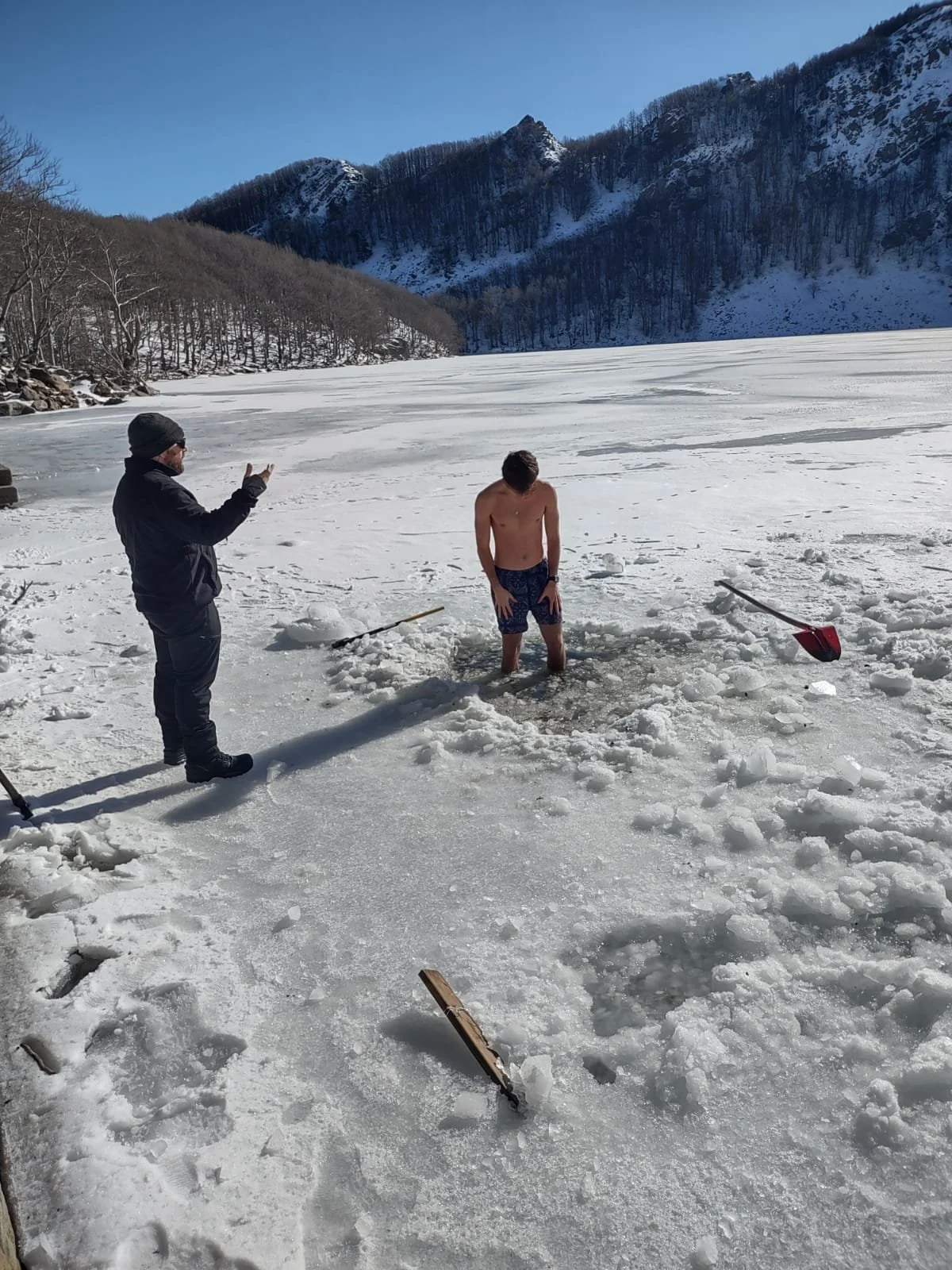 Una persona senza maglietta sta immersa in un lago ghiacciato, circondata da neve e montagne.
