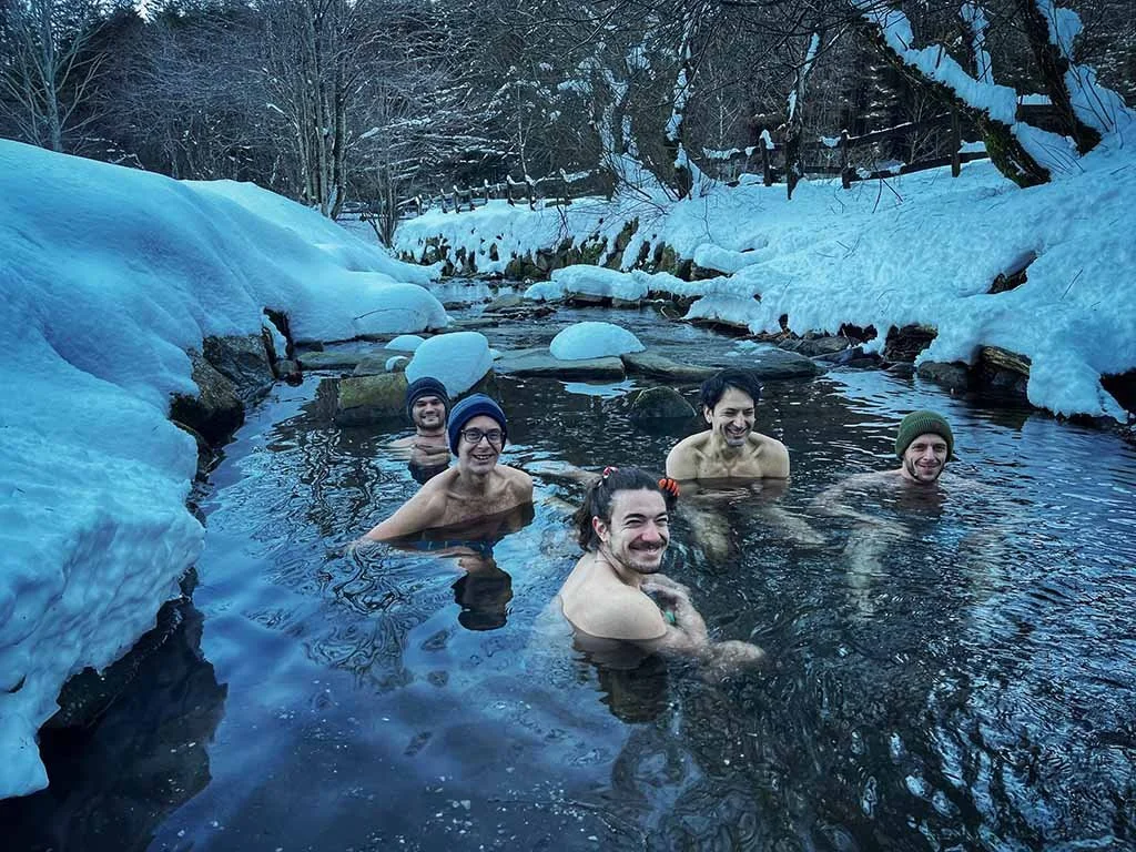 Gruppo di persone che fanno il bagno in una sorgente termale all'aperto circondata da neve e alberi in inverno.