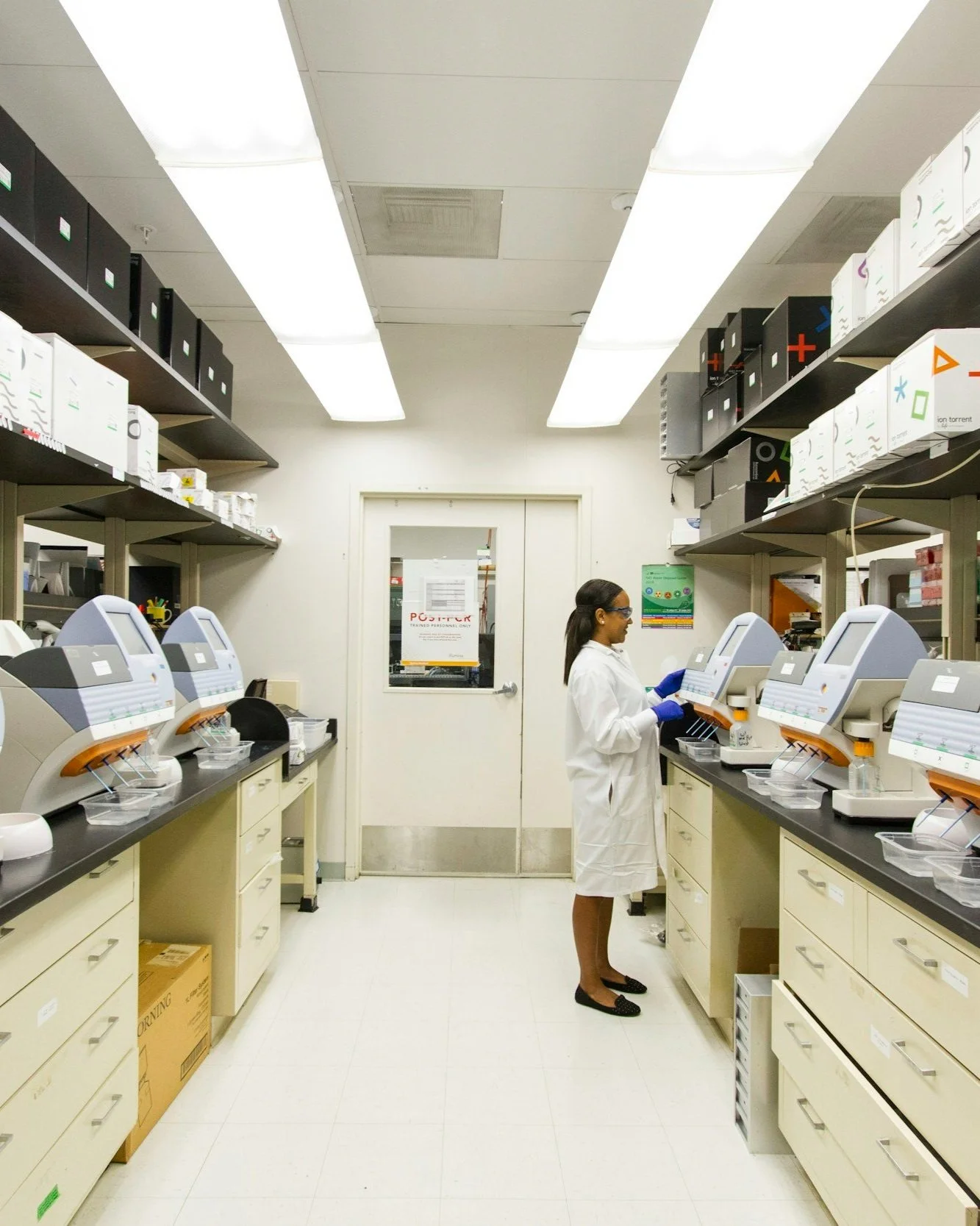 A scientist in a white lab coat and blue gloves working with laboratory equipment in a lab with white walls and ceiling, fluorescent lighting, shelves with boxes and equipment, and a closed door at the back.