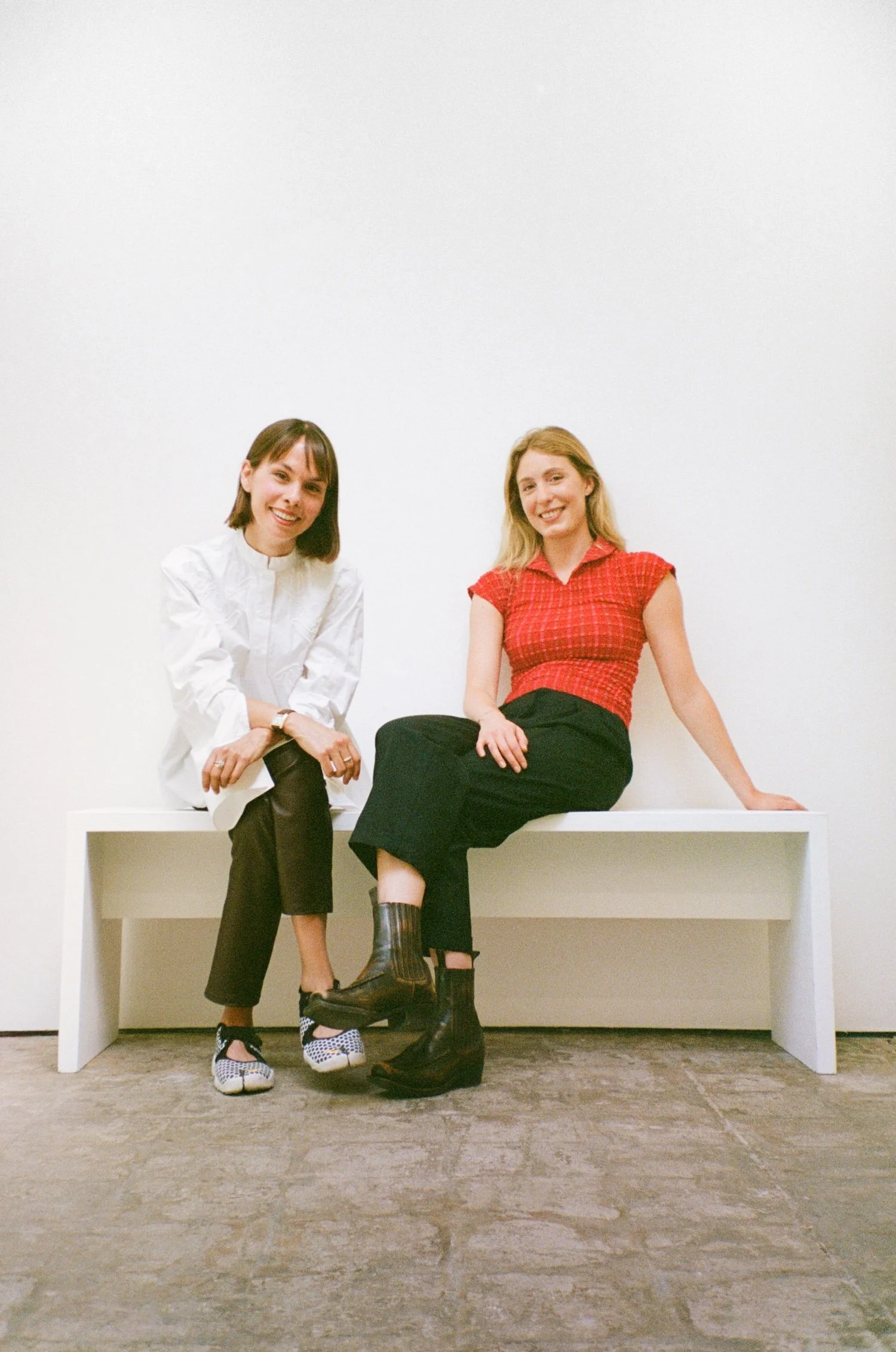 Two women sitting on a white bench against a plain white wall, smiling at the camera.