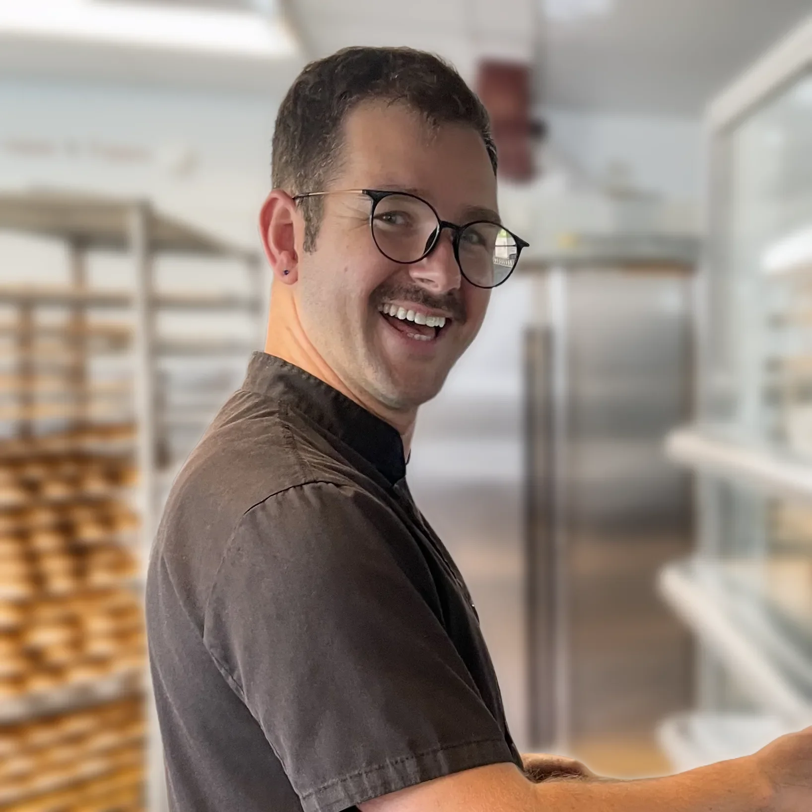 Happy, smiling man wearing glasses in black chefs shirt, with head turned toward camera