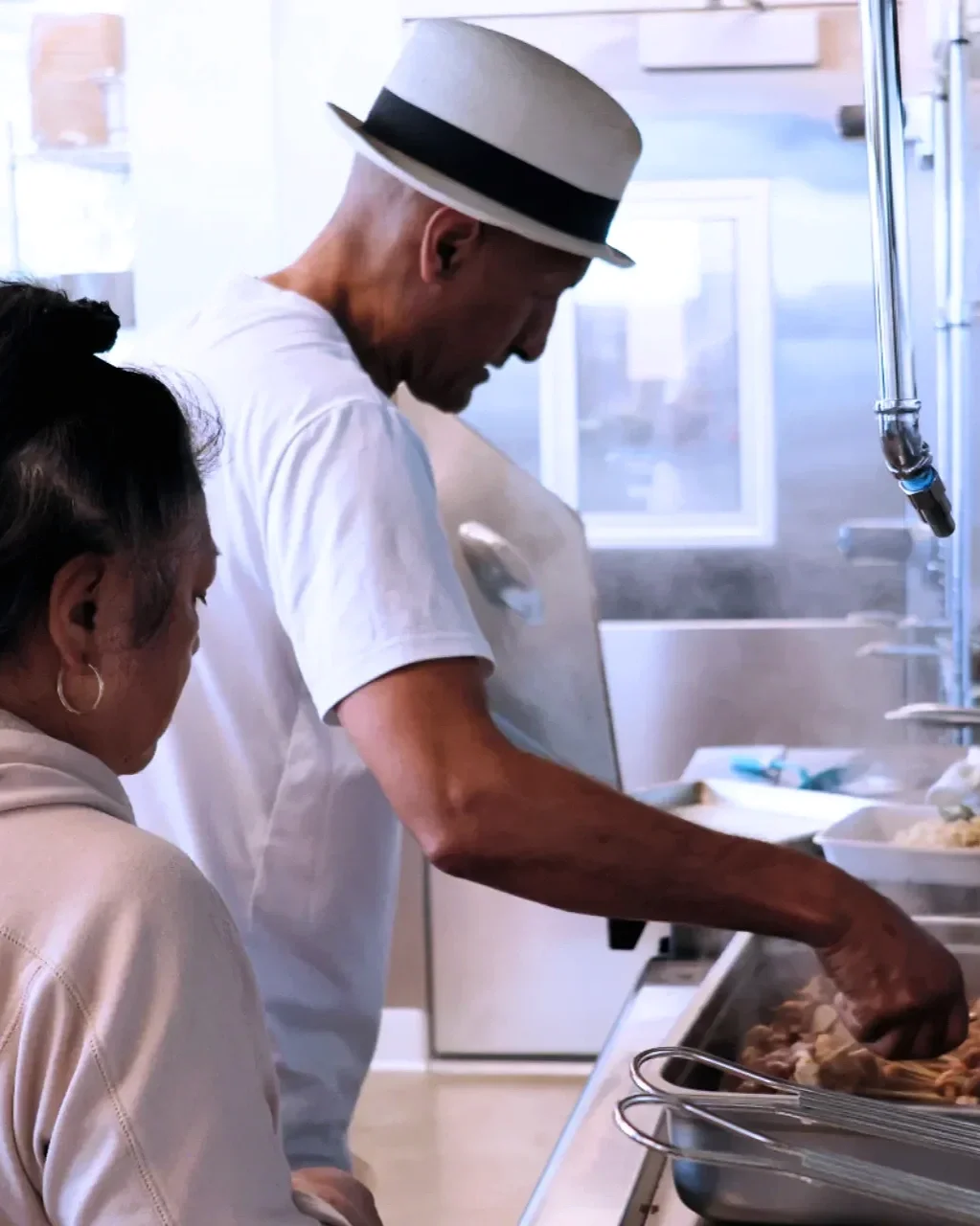 A man wearing a fedora hat and white t-shirt preparing food in a kitchen, with a woman nearby.