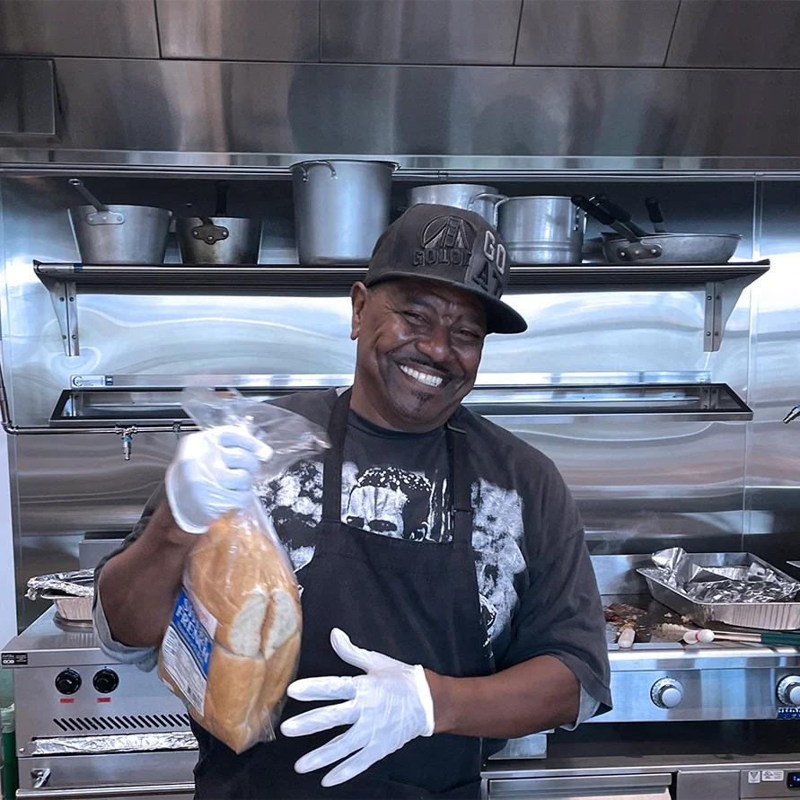 Smiling man in black t-shirt, baseball cap, and black apron with face toward camera, holding a package of bread