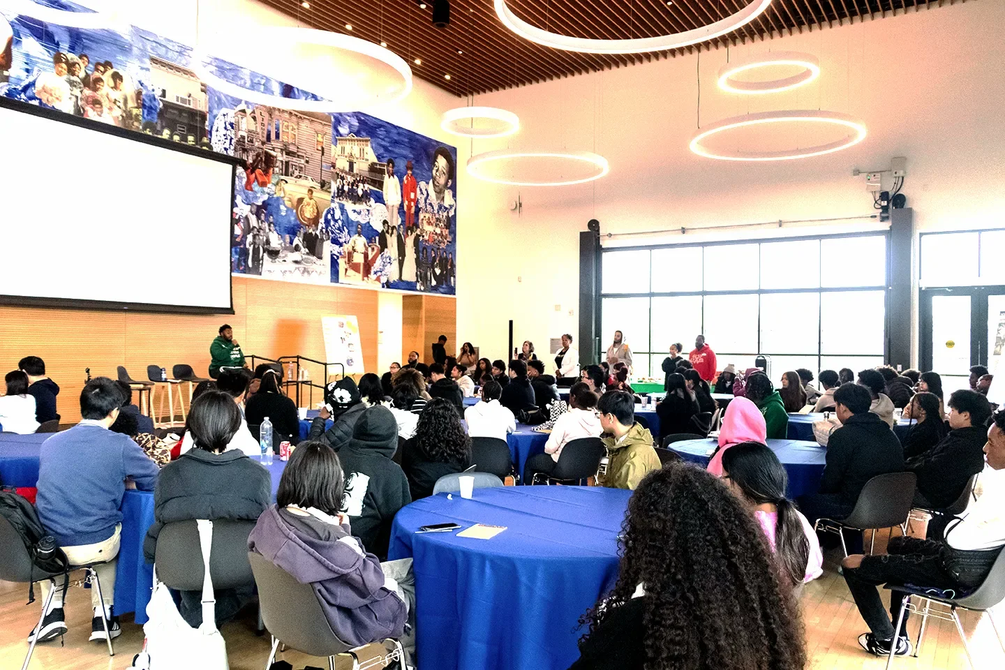 Groups of people seated at tables in a large auditorium listening to a speaker