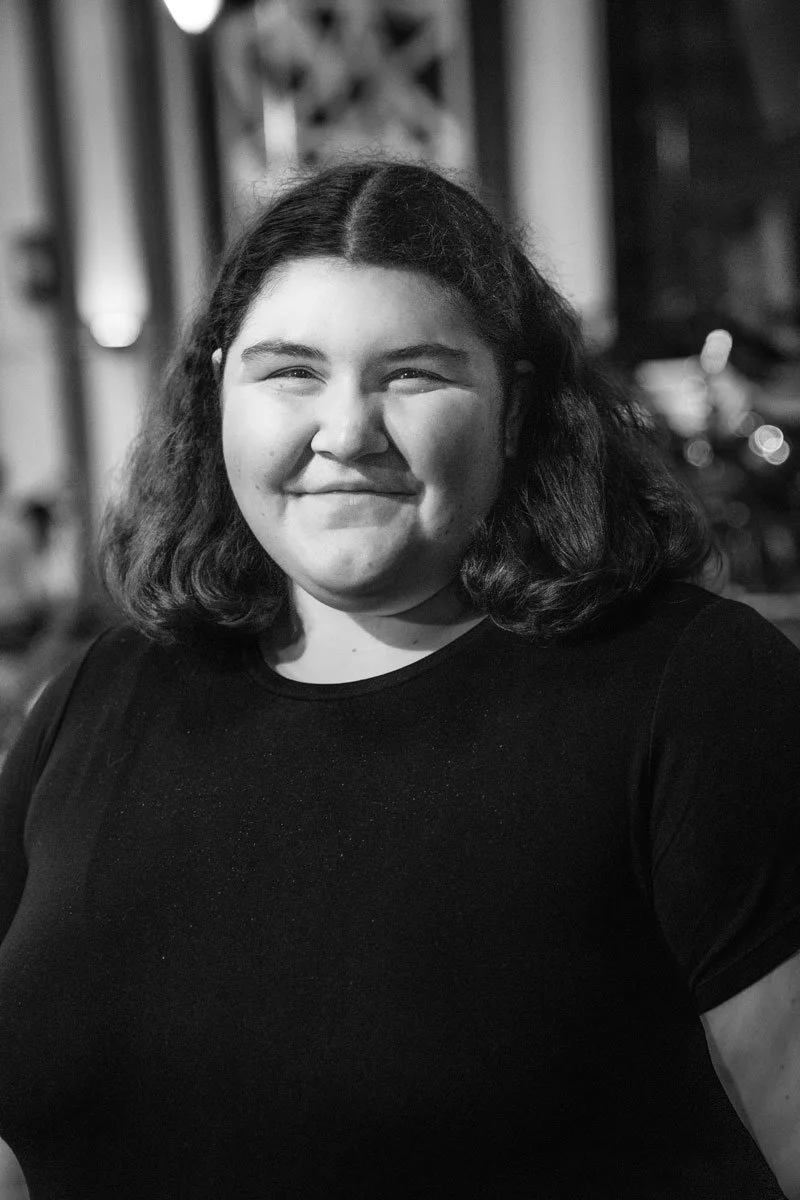 A young woman with shoulder-length wavy hair smiling at the camera, wearing a black shirt, in an outdoor nighttime setting.