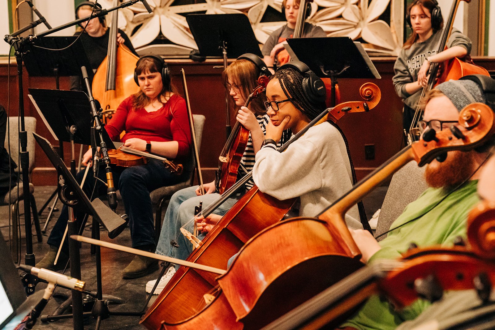 Orchestra musicians in a recording studio, including a woman with glasses and braided hair playing the cello, and other musicians with violins, a guitar, and a double bass, all wearing headphones.