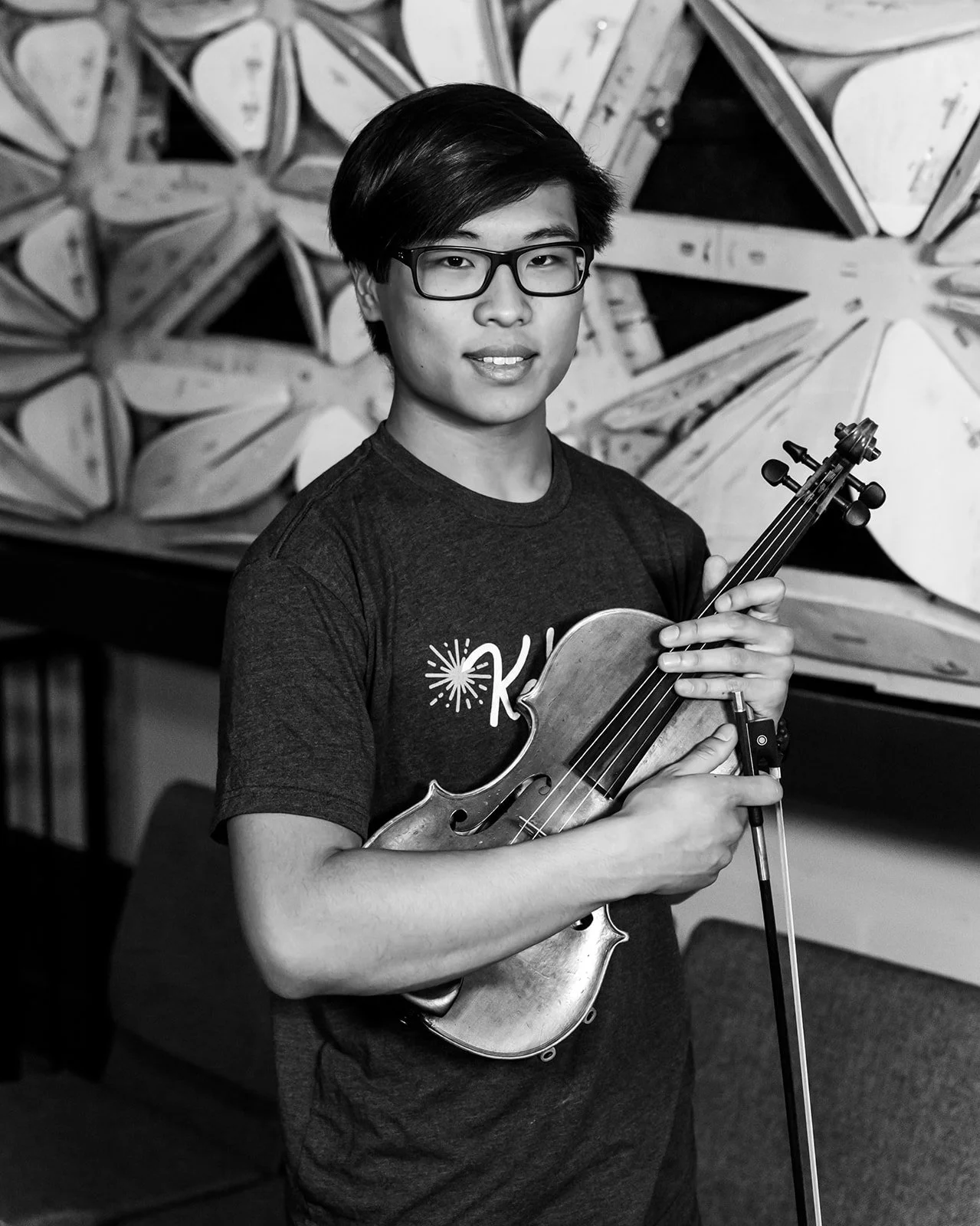 A young person with glasses holding a violin and bow, standing in front of a geometric, abstract wall art.