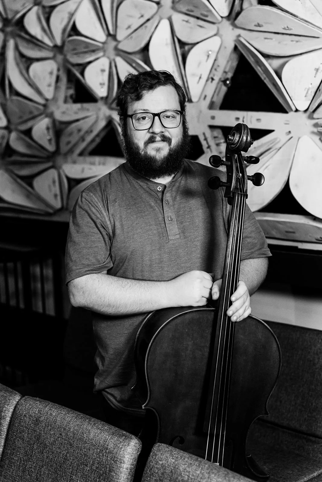 A man with glasses and a beard holding a cello, standing in front of a wall with art pieces in an indoor setting.