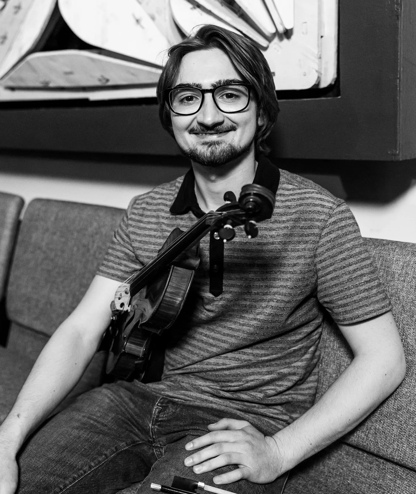 A young man with glasses and a striped T-shirt sitting on a couch, holding a violin with a bow resting on his shoulder, smiling at the camera in black and white.