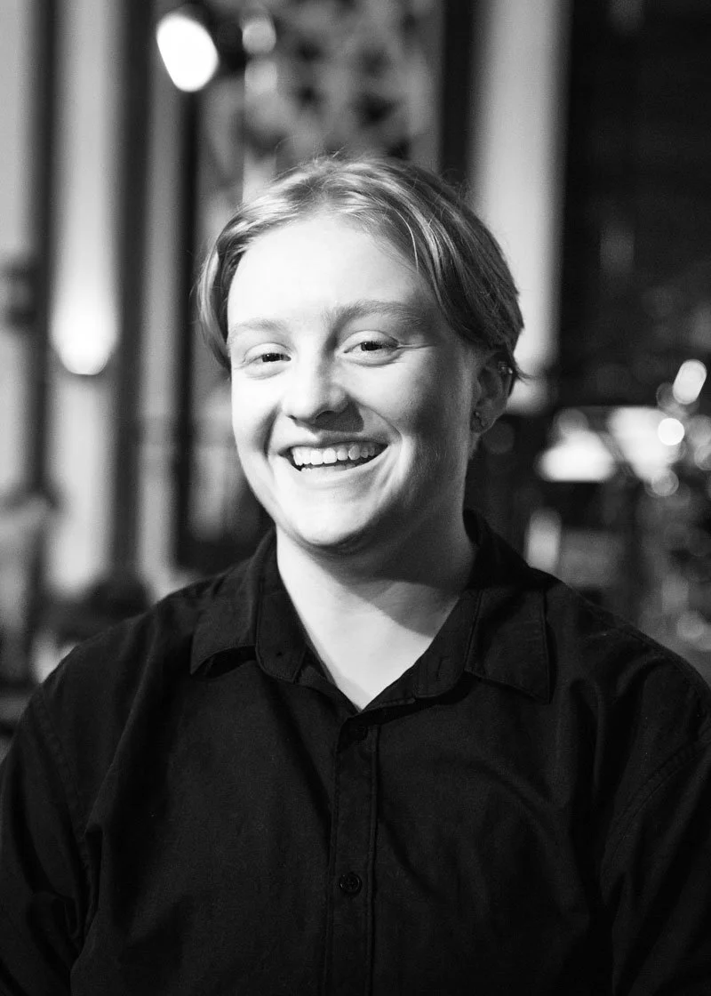 A smiling young woman with short hair wearing a dark button-up shirt, sitting indoors with a blurred background.