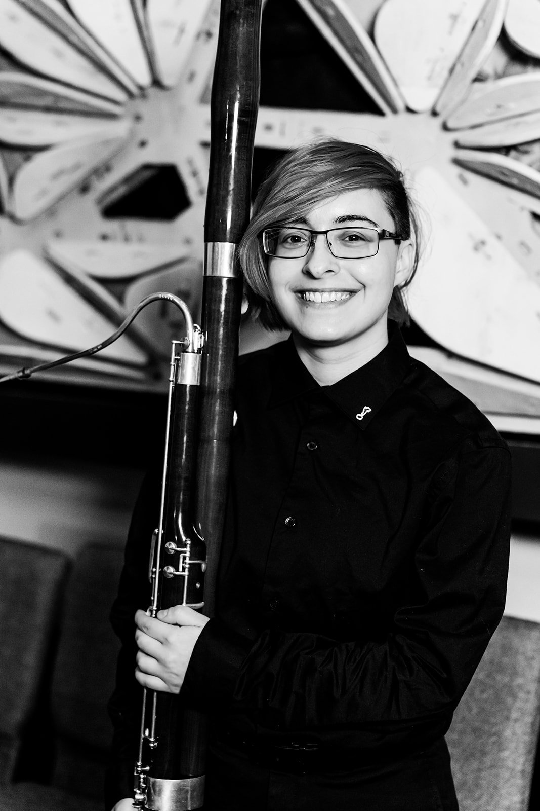 A young woman with short hair and glasses smiling, holding a musical instrument, standing in front of a butterfly-shaped background.