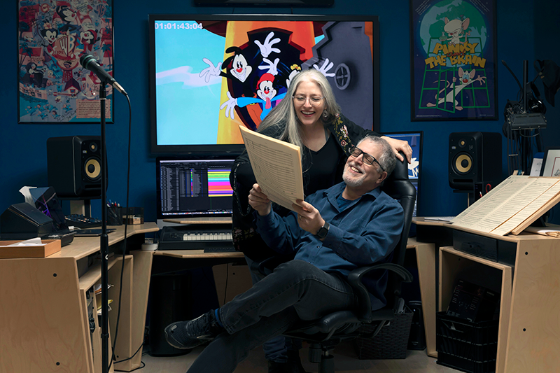 Two people, a woman and a man, are happily working together in a recording studio filled with music equipment, computers, and posters. The woman stands behind the seated man, who is reading a script or sheet music, both smiling. A large screen behind them displays animated characters, and there are speakers and workstations around them.