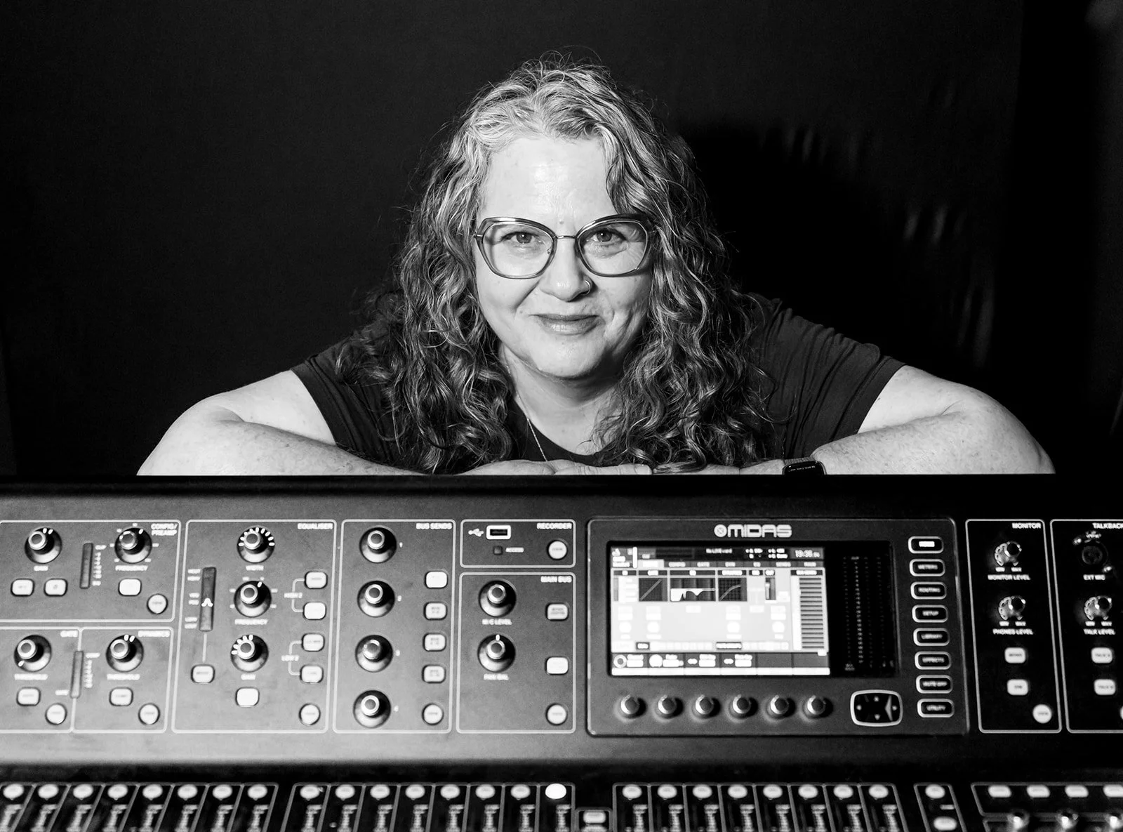 Black and white photo of a woman with curly hair and glasses, resting on her arms on a large electronic music mixing console or synthesizer, with a dark background.