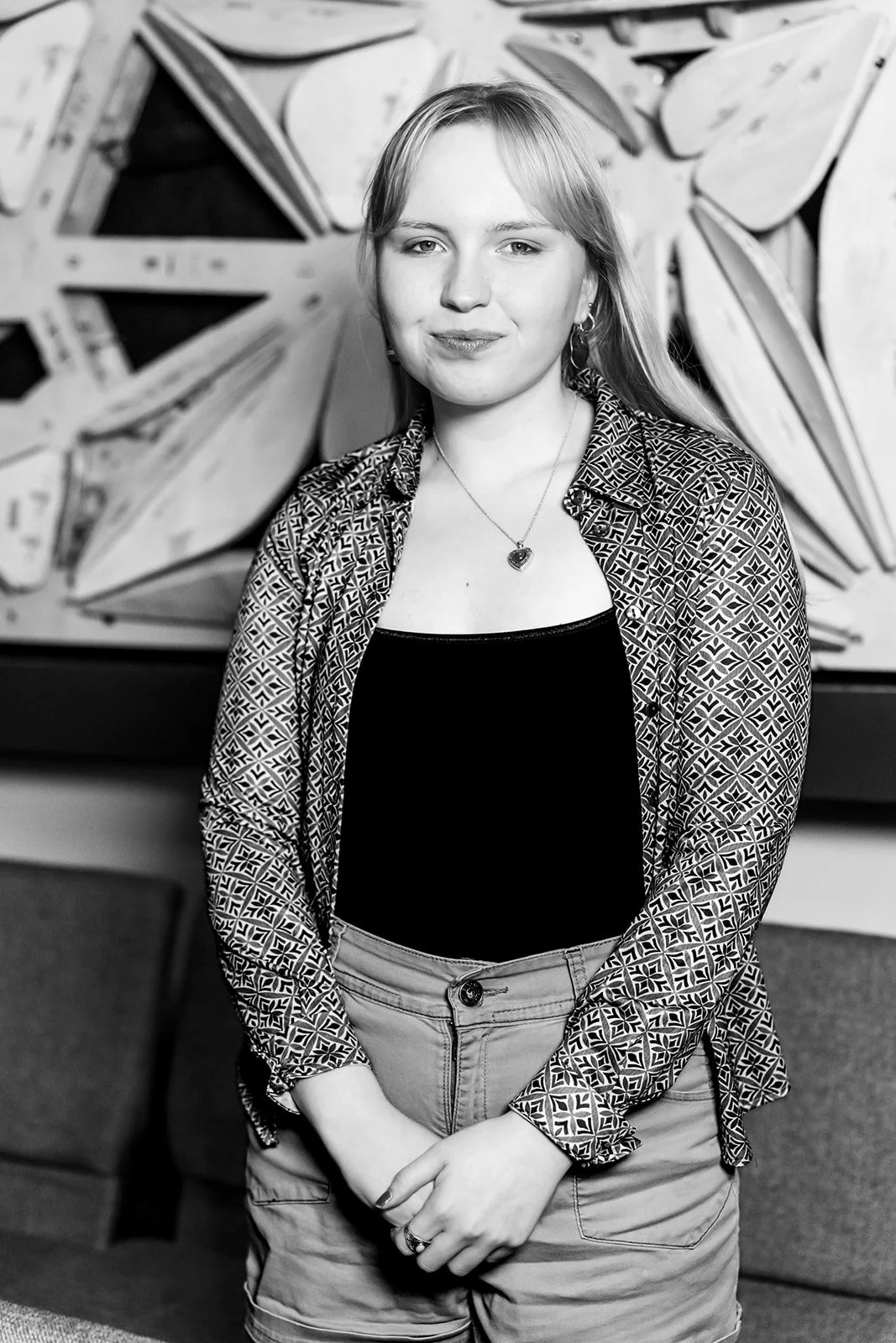 A young woman with light hair, wearing a patterned shirt, black top, and cargo shorts, standing in front of a large abstract artwork in black and white.