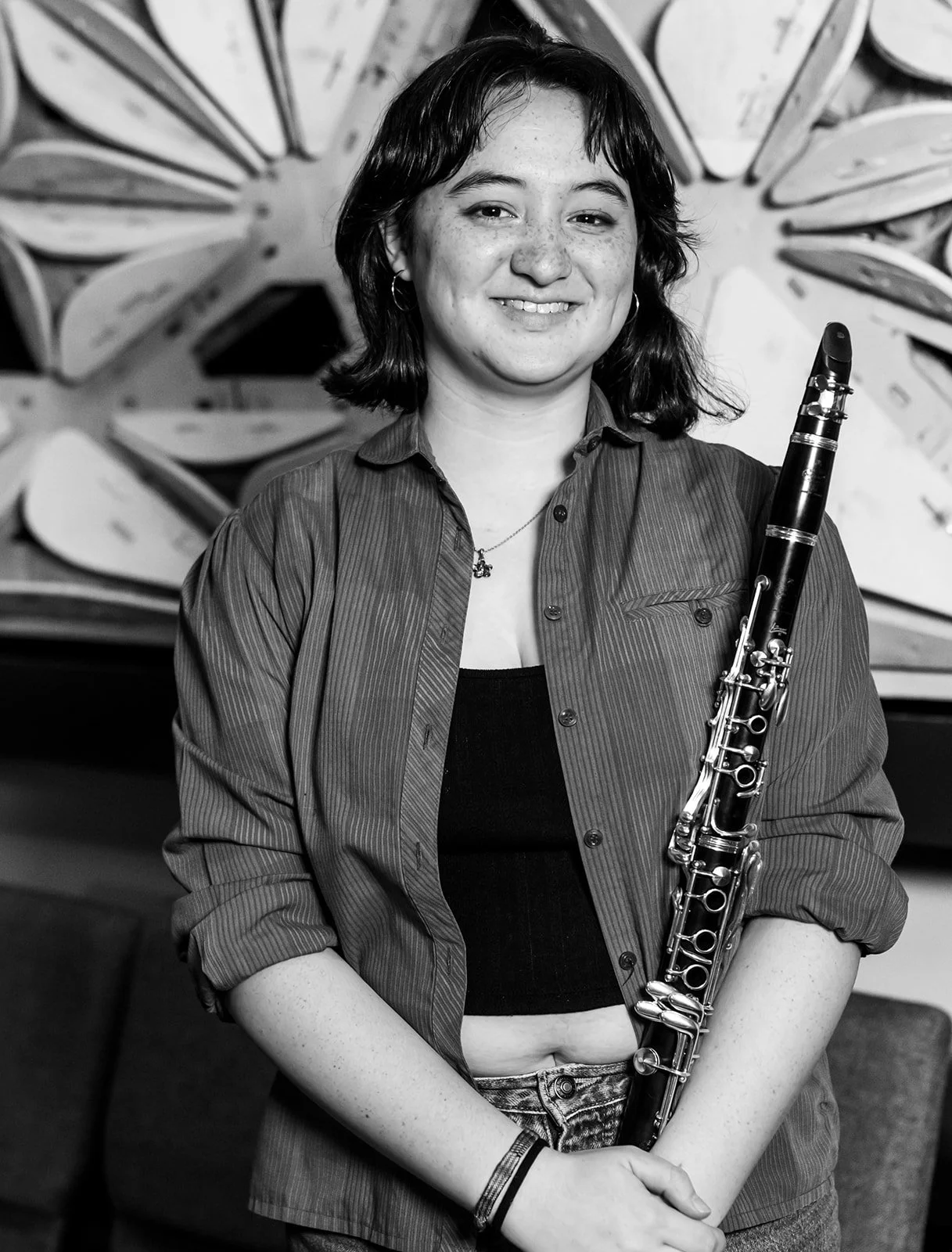 Black and white photo of a young woman with short dark hair, smiling, holding a bassoon, standing in front of wooden sculpture art.