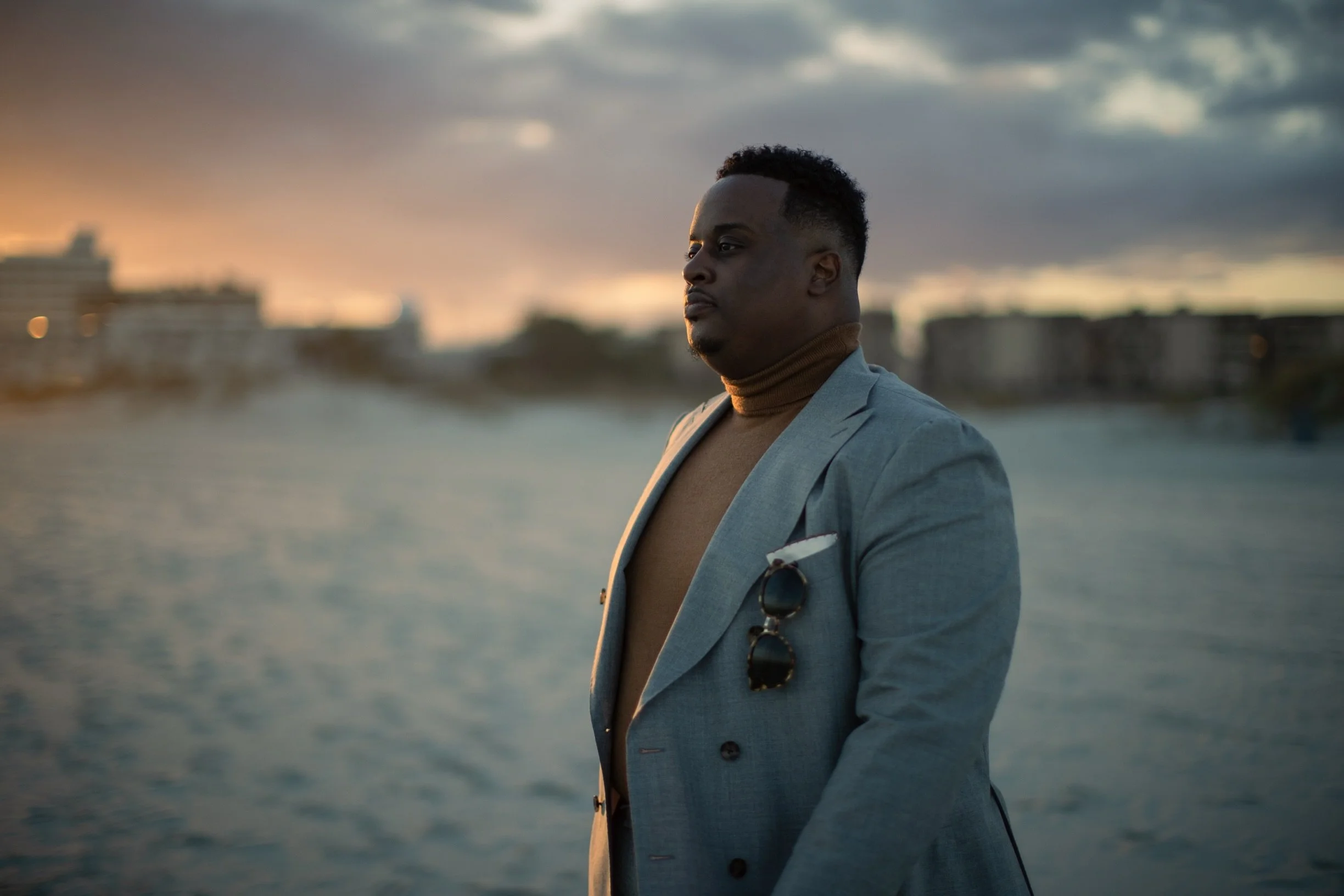 A man in a gray blazer and tan turtleneck stands by a body of water during sunset, with city buildings blurred in the background.