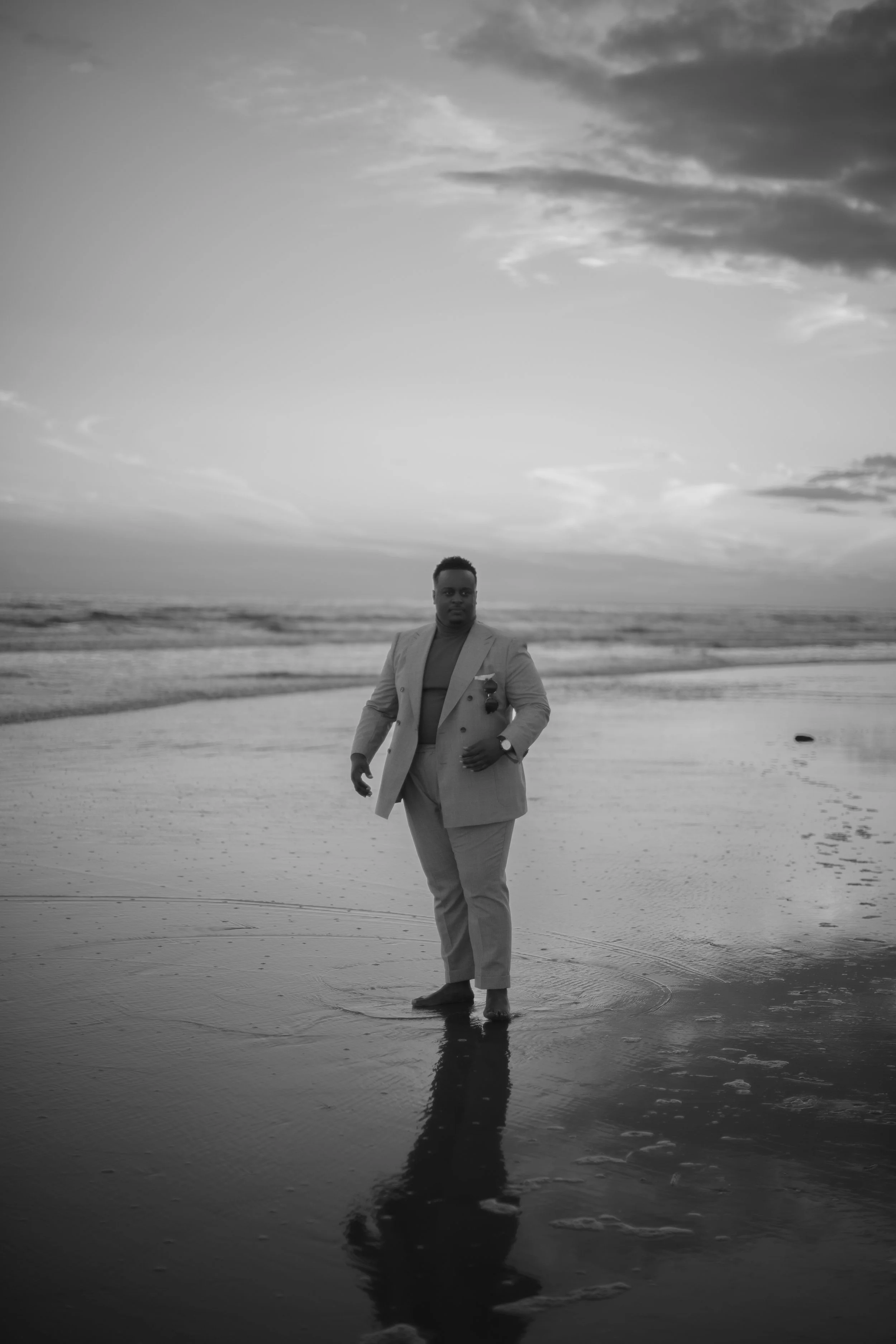 A man dressed in a suit stands barefoot on the beach with his reflection visible in the wet sand, during sunset or sunrise.