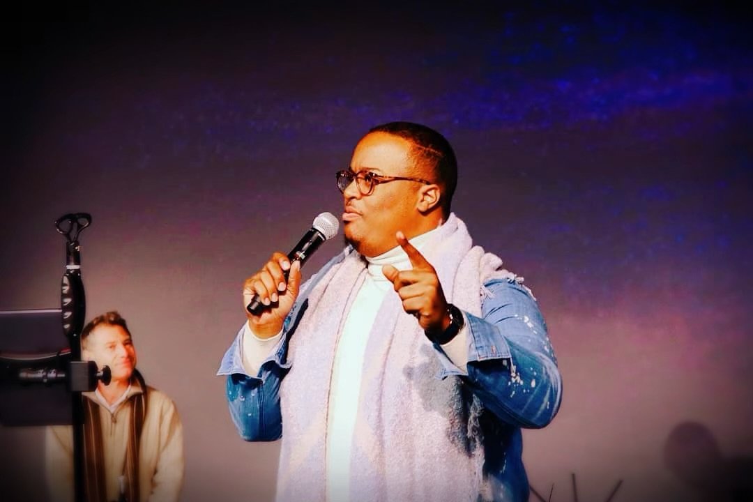 A man in glasses holding a microphone, pointing with his finger, with a woman in the background looking at him, against a dark blue and purple sky.