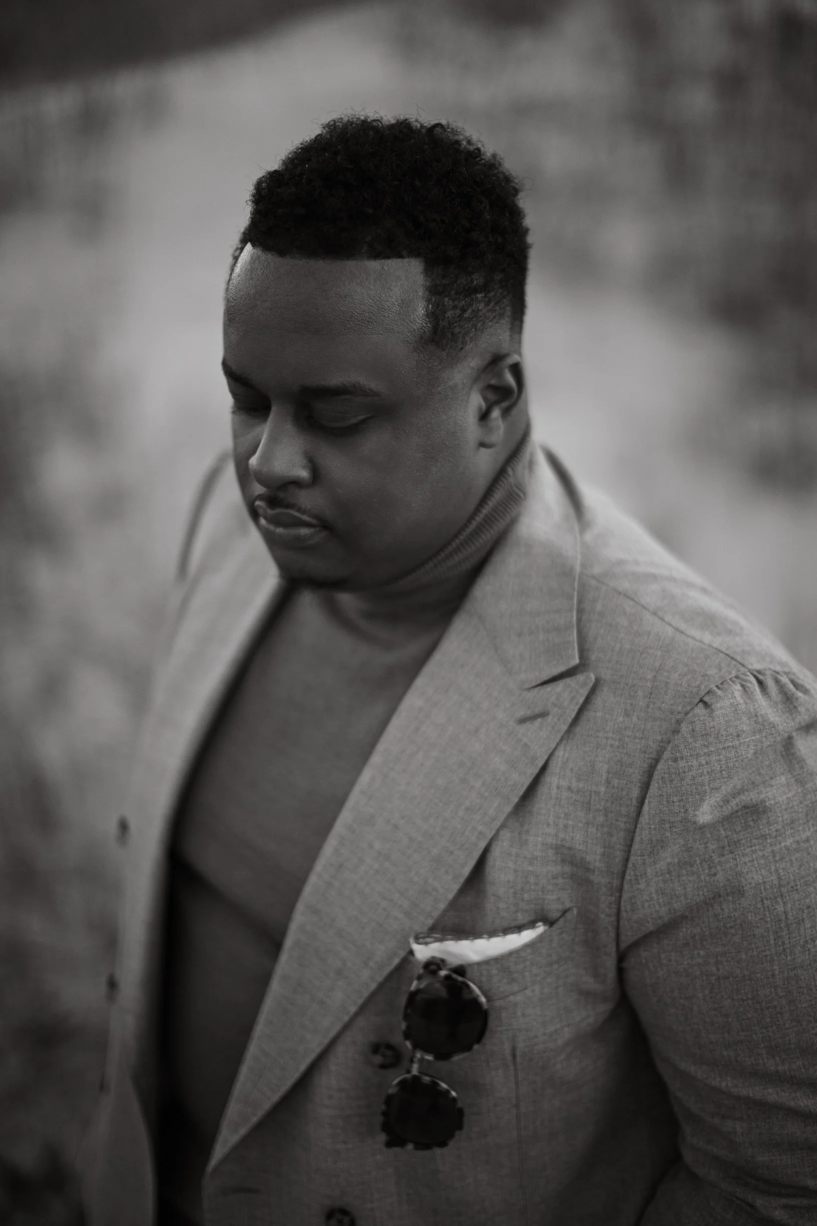 A black and white portrait of a man with short, curly hair, wearing a light-colored blazer and turtleneck, with sunglasses tucked into the blazer's pocket.