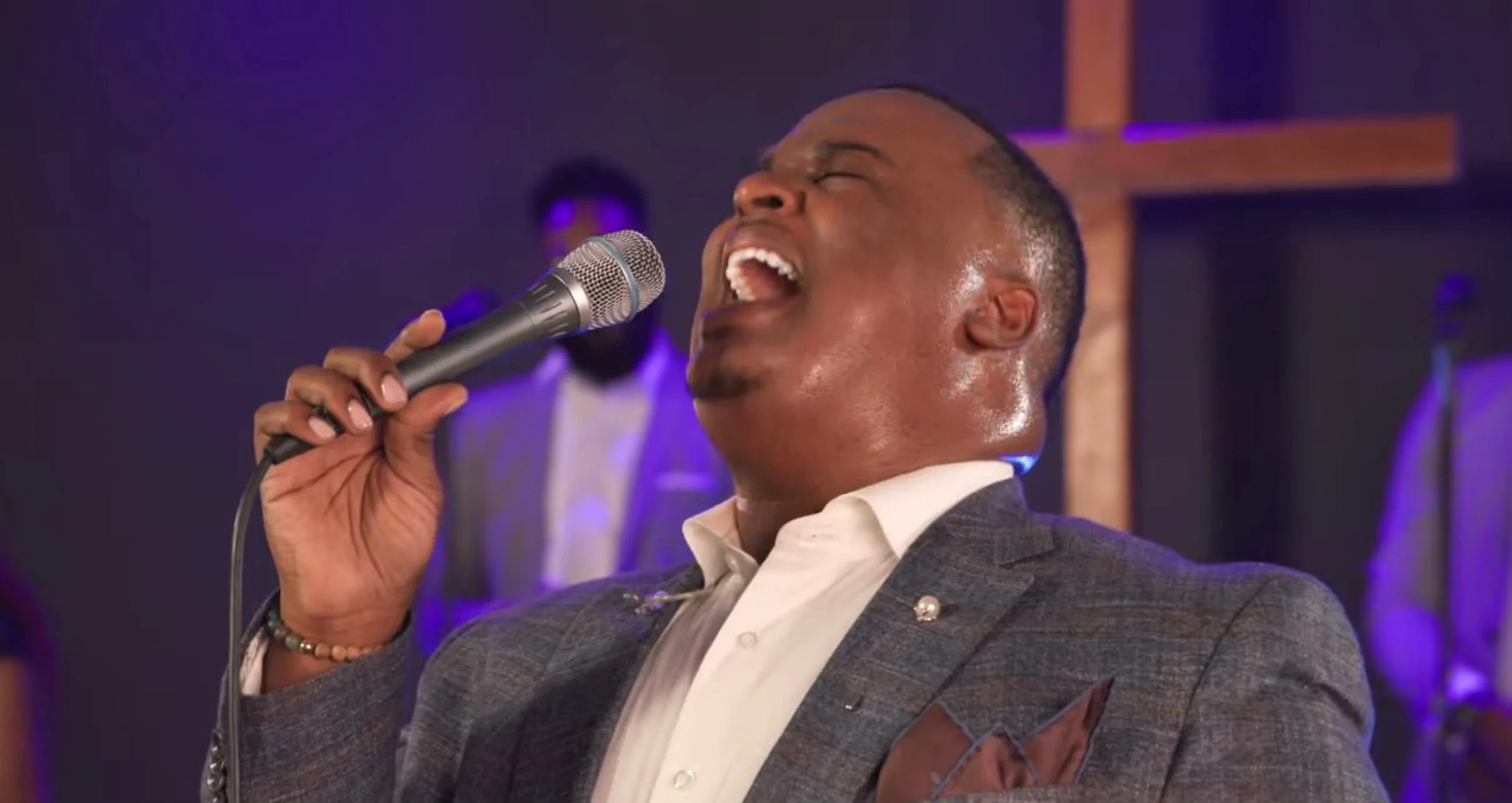 Man singing passionately into a microphone in a church or religious setting with a wooden cross in the background.