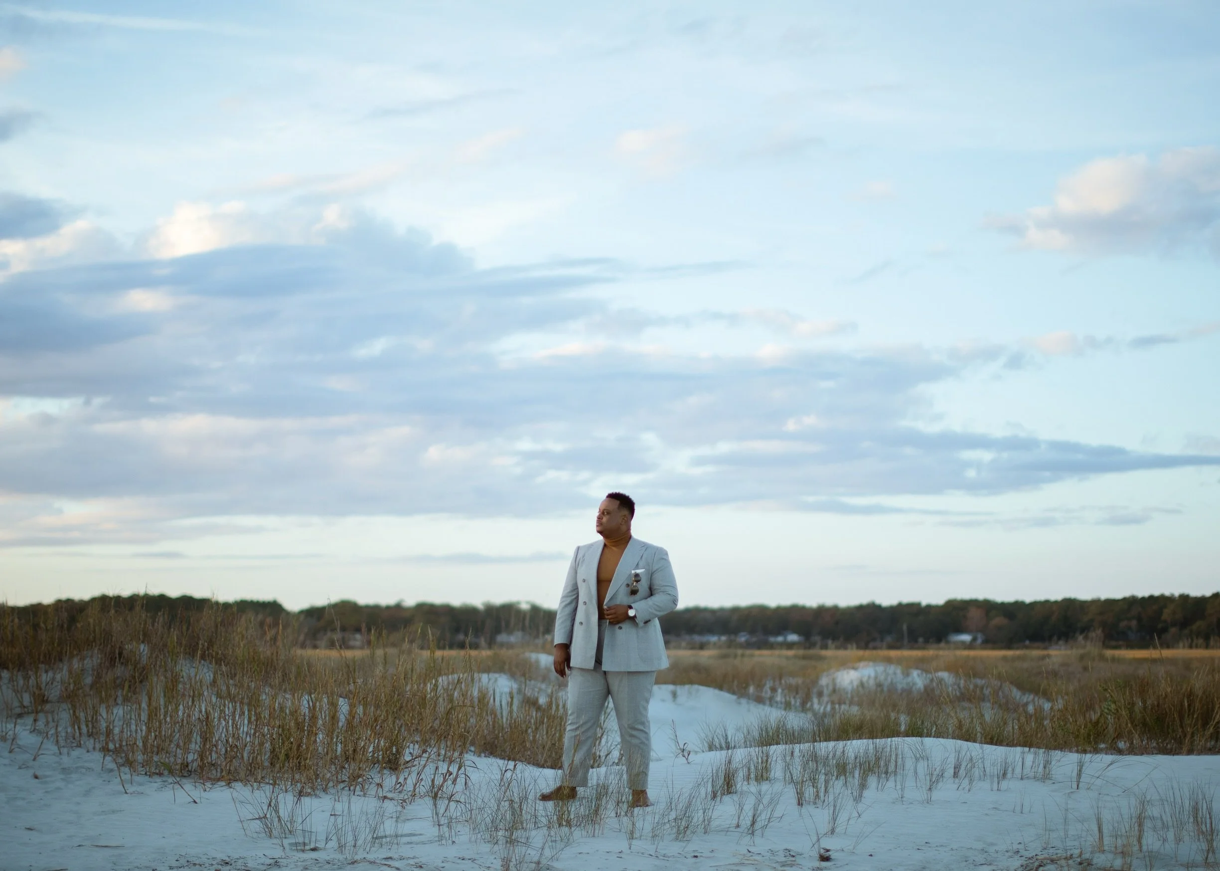 A man dressed in a light gray suit stands on sandy dunes with dry grass in a flat landscape, under a blue sky with scattered clouds.