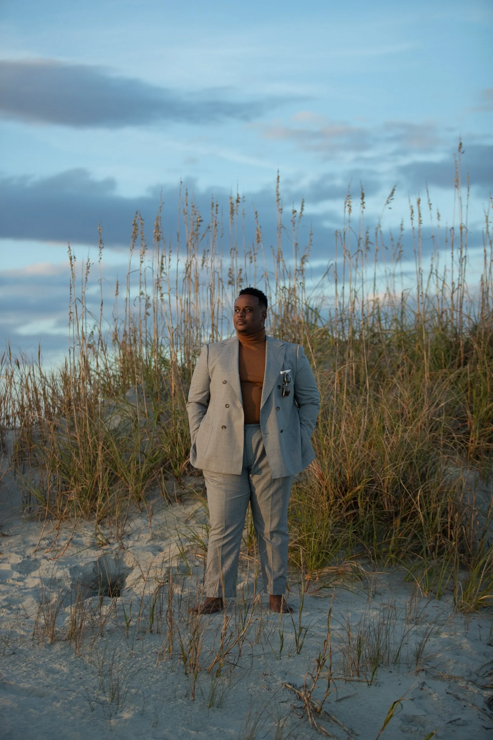 A man in a gray suit standing barefoot on a sandy beach with tall grass, during sunset or sunrise with a blue sky and clouds in the background.