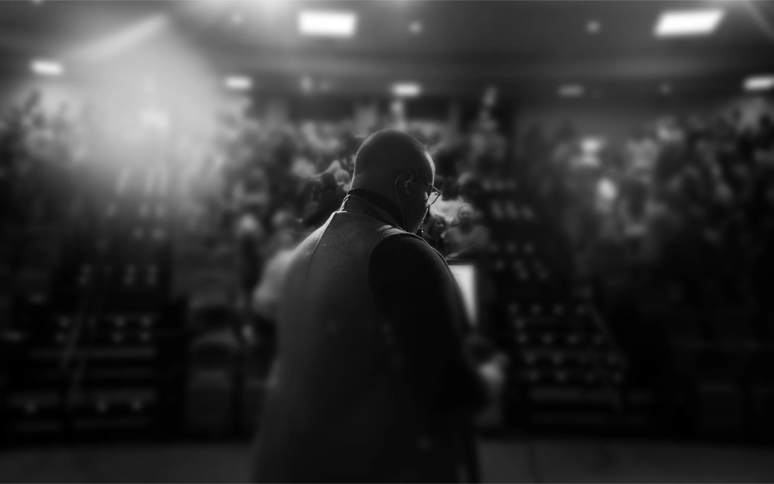 A man in glasses and a mask, seen in profile, standing in a packed auditorium with rows of chairs and people in the background.