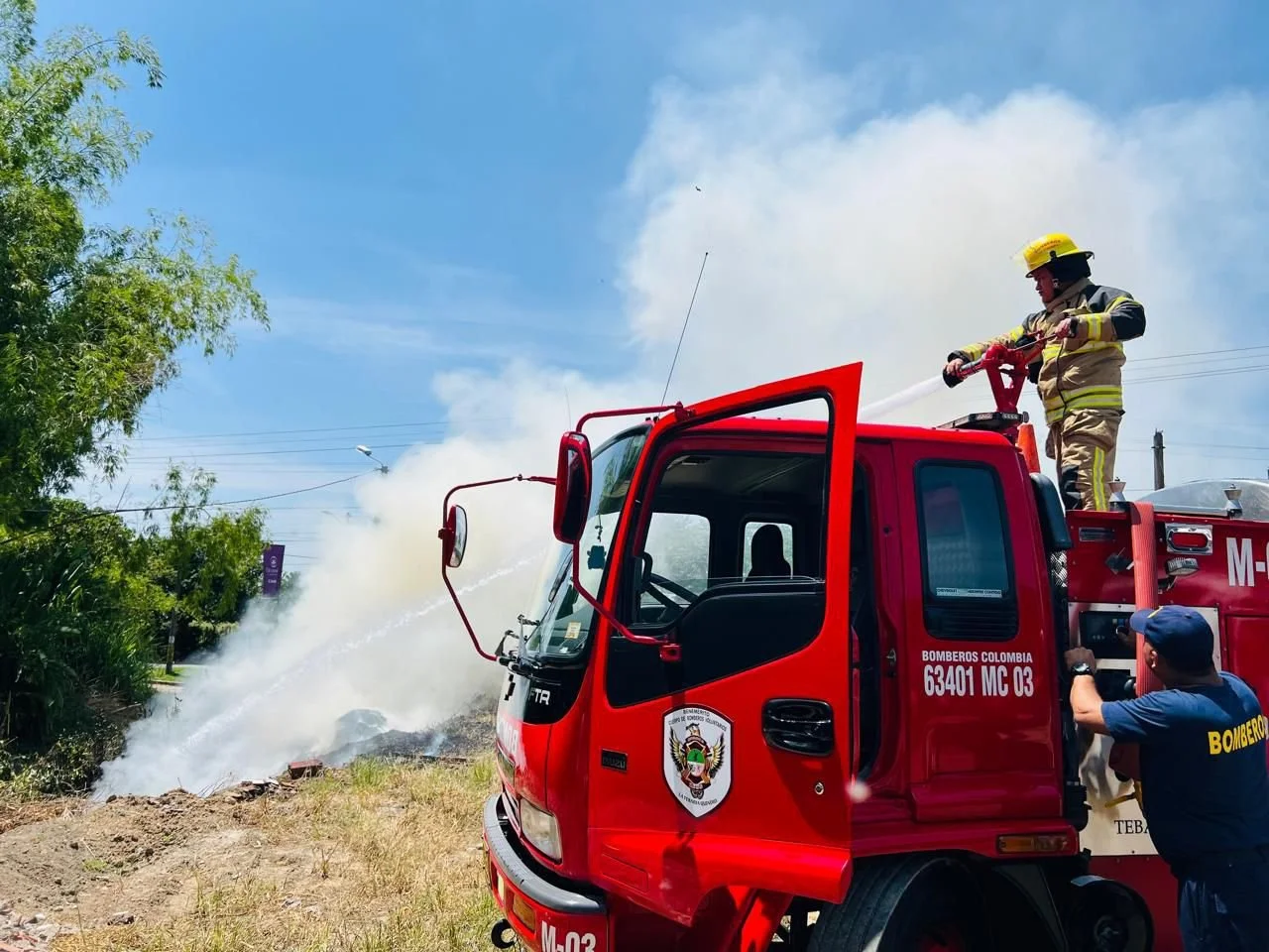 Bomberos colombianos combaten un incendio con mangueras en un camión de bomberos rojo, mientras uno de ellos está en la parte superior con equipo de protección personal y otro en el suelo ayuda a supervisar la situación con vestimenta de bombero.