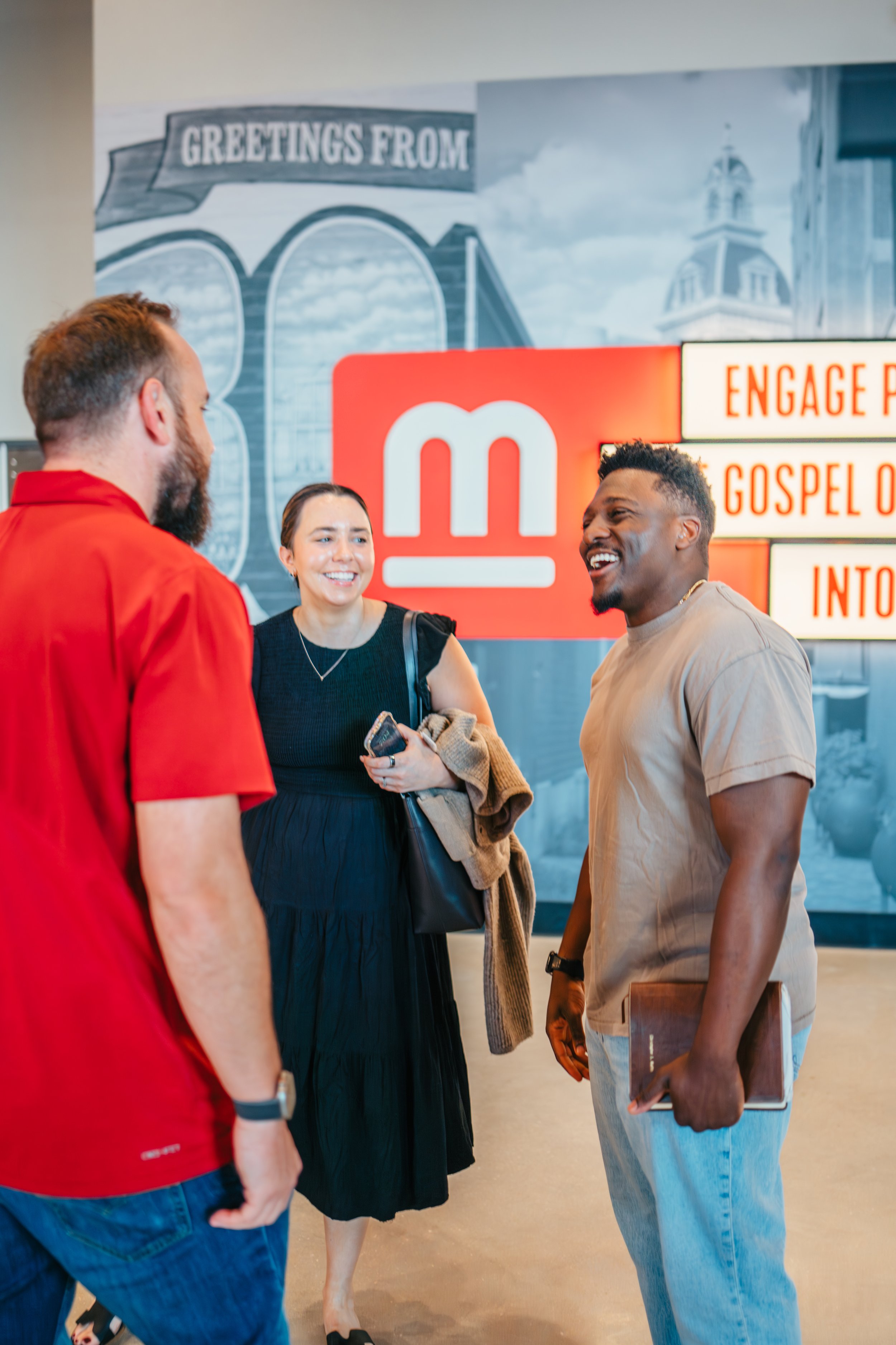 Three people having a conversation in front of a red sign with the letter 'm' and partially visible words. The background shows a mural with a bridge and buildings, and the setting appears to be a public indoor space.