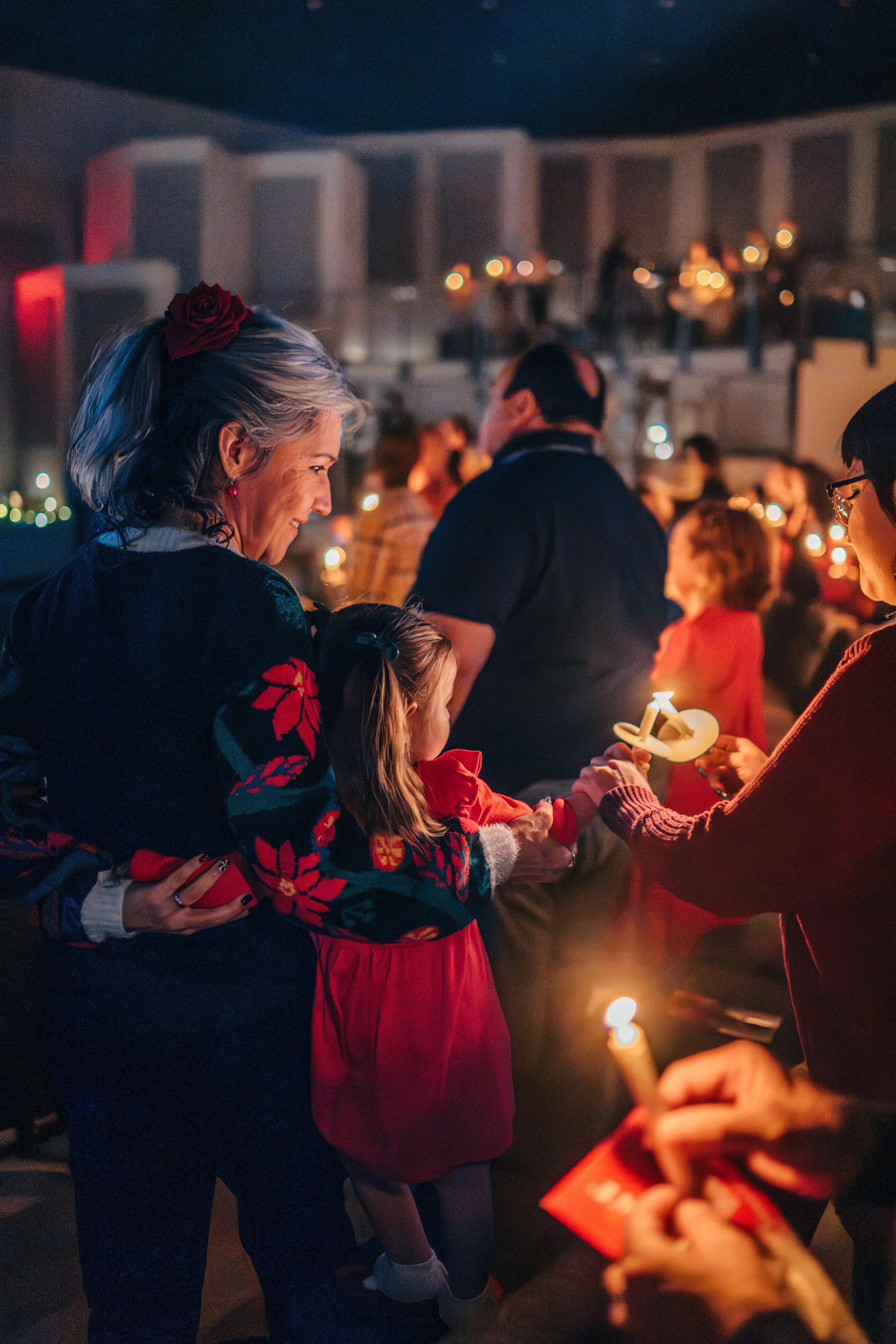 People participating in a candle lighting ceremony during a Christmas event, with children and adults holding lit candles amid festive decorations and Christmas sweaters.