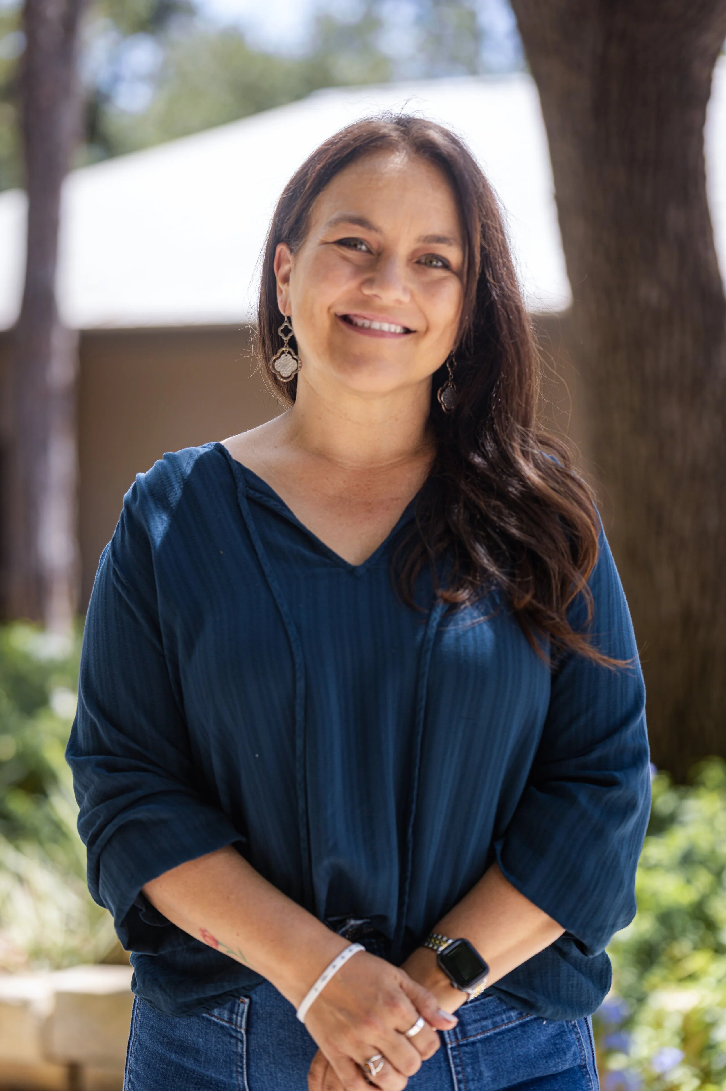 A woman with long brown hair and light skin, smiling, wearing a navy blue top and earrings, standing outdoors near trees and a house.