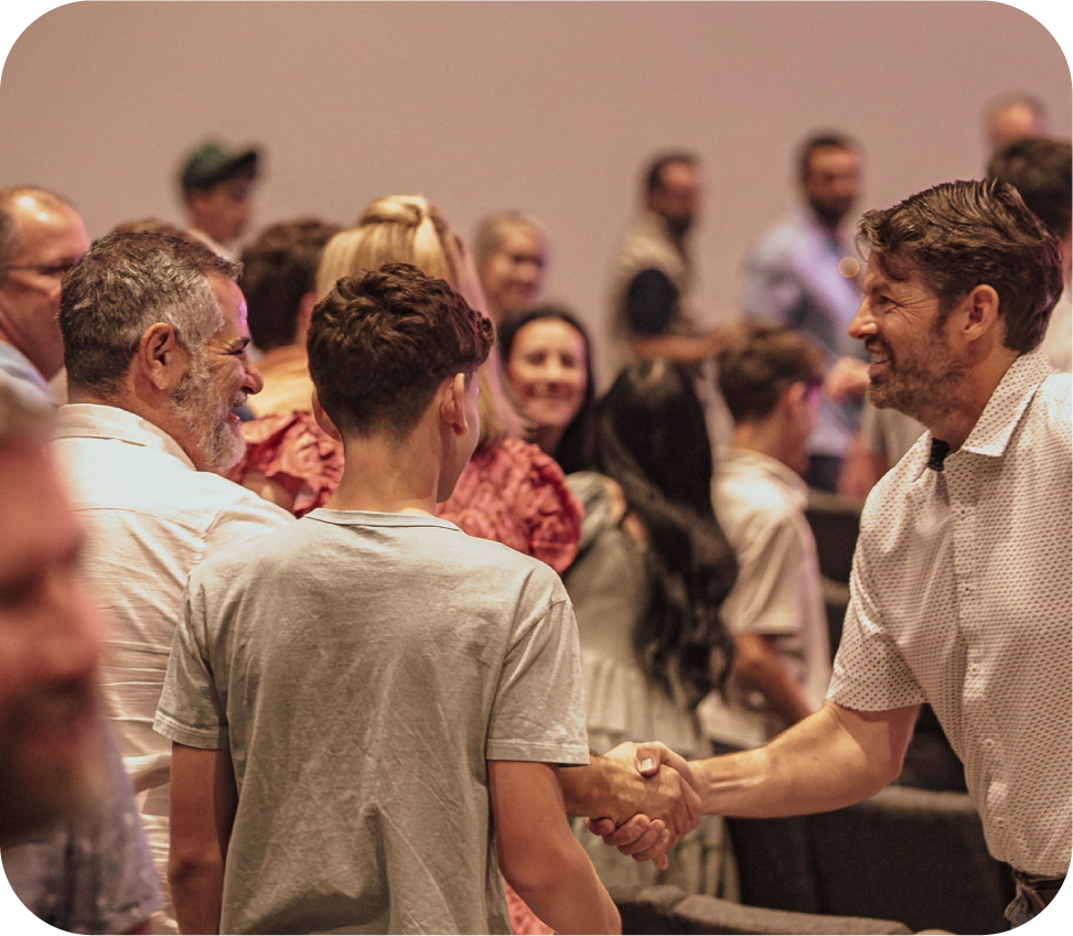 Two men shaking hands in a crowded men's event, with several people smiling and observing in the background.