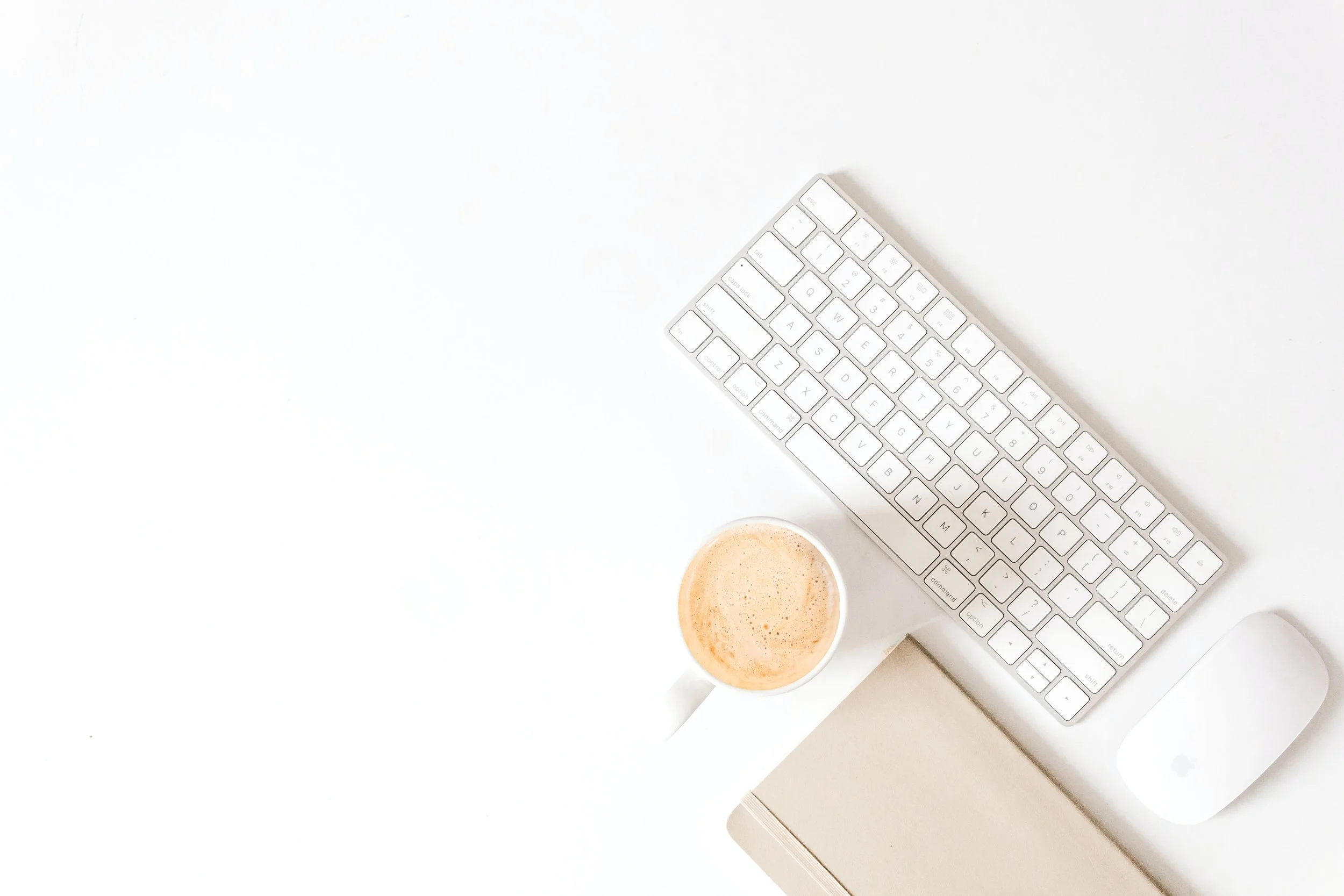 A white desk setup including a wireless keyboard, a wireless mouse, a closed beige notebook, a cup of coffee with foam, and a light-colored background.