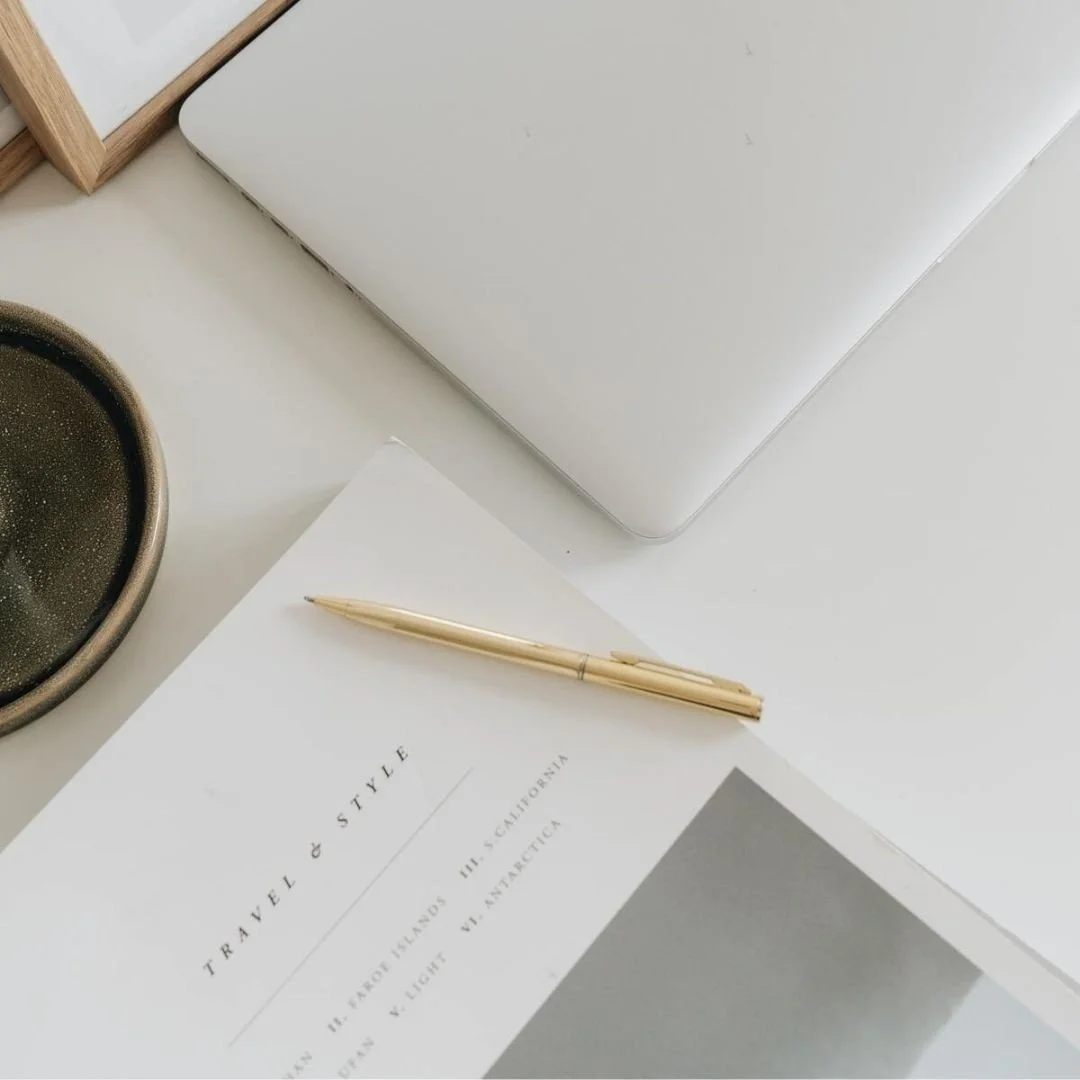 A white desk with a closed silver laptop, a white sheet of paper with text, a gold pen, a black textured bowl, and a wooden picture frame.