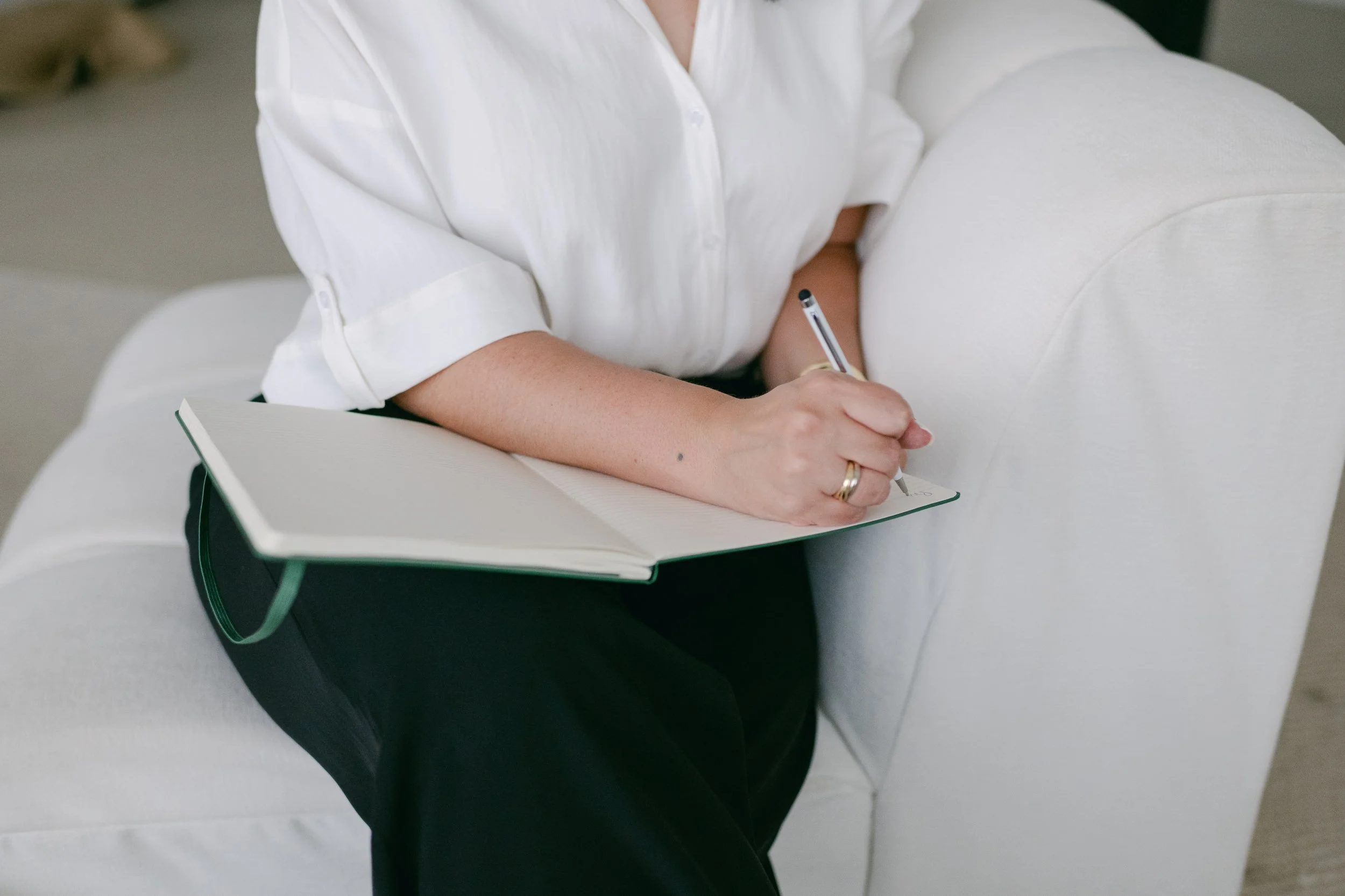 Person sitting on a white sofa, writing in a notebook with a pen, wearing a white shirt and black pants.