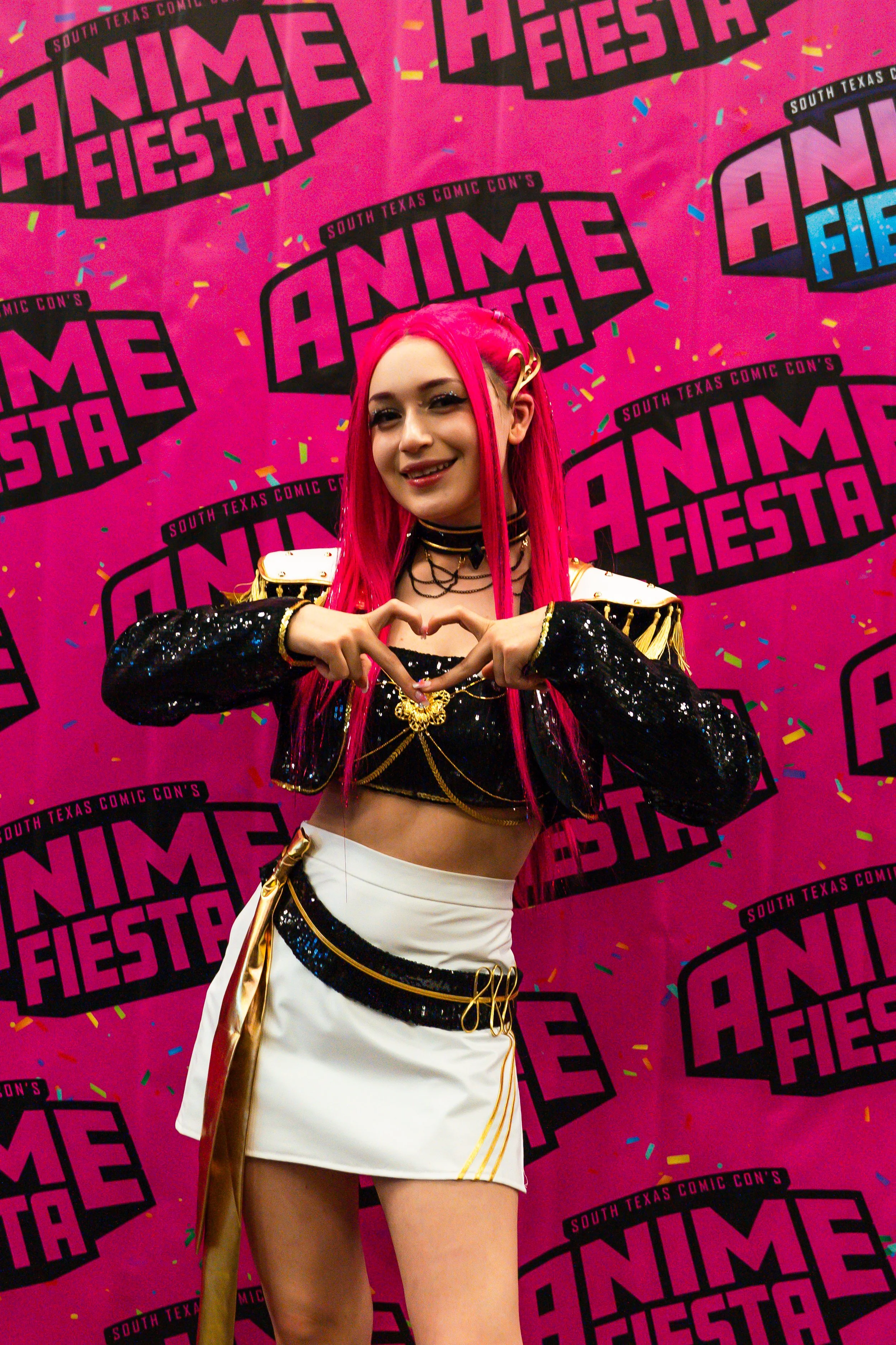 A person with long pink hair dressed in a black and white costume with gold accents, making a heart shape with their hands, at South Texas Comic Con's Anime Fiesta event.