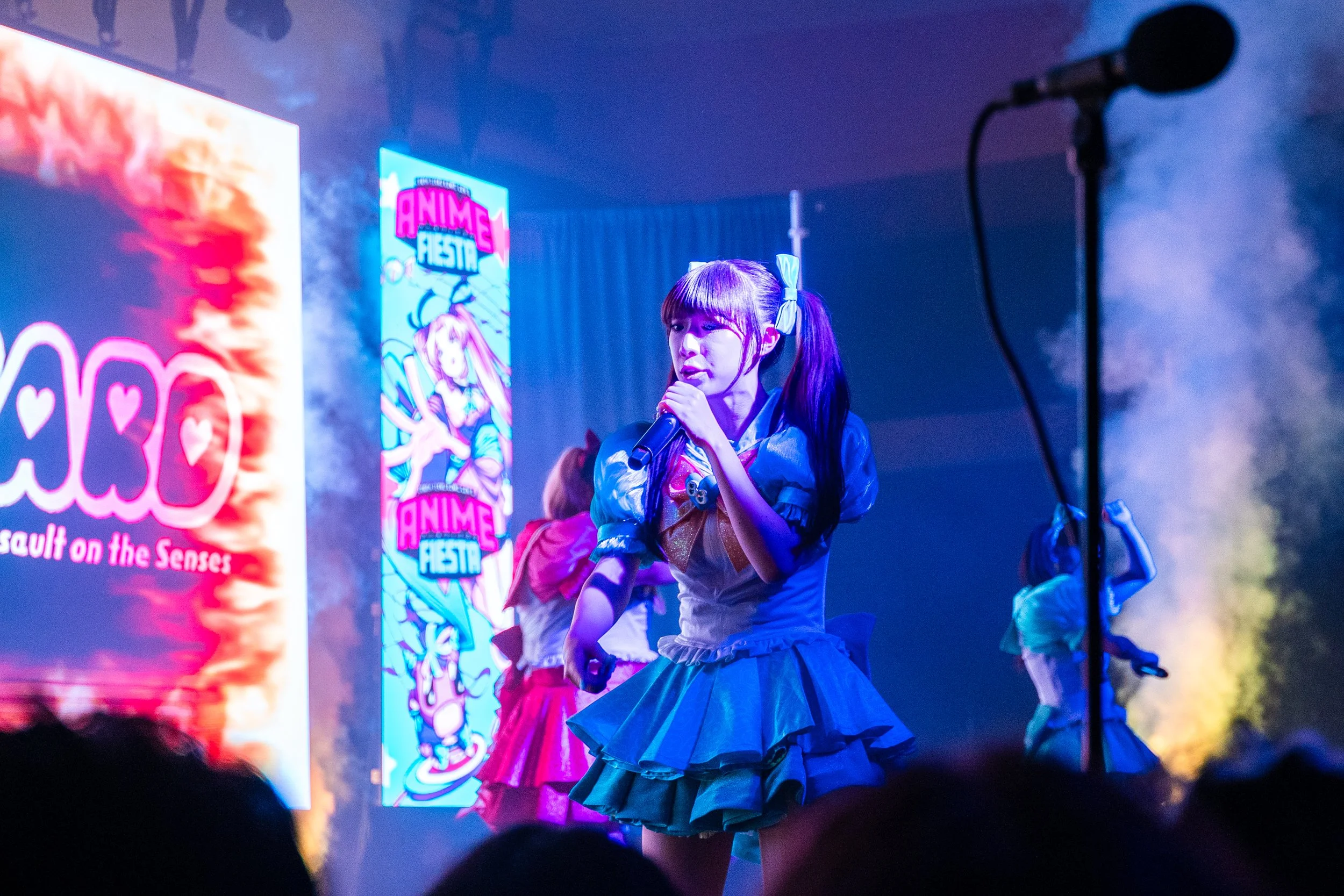 A young woman dressed in a colorful costume performs on stage at an anime festival, holding a microphone with vibrant illuminated signage in the background.