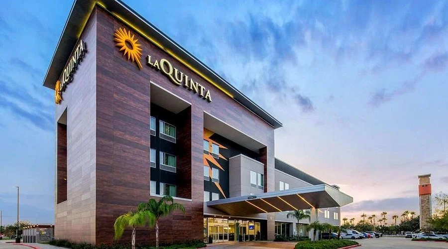 Exterior of La Quinta hotel with modern architecture, palm trees, parking lot, and a partly cloudy sky.