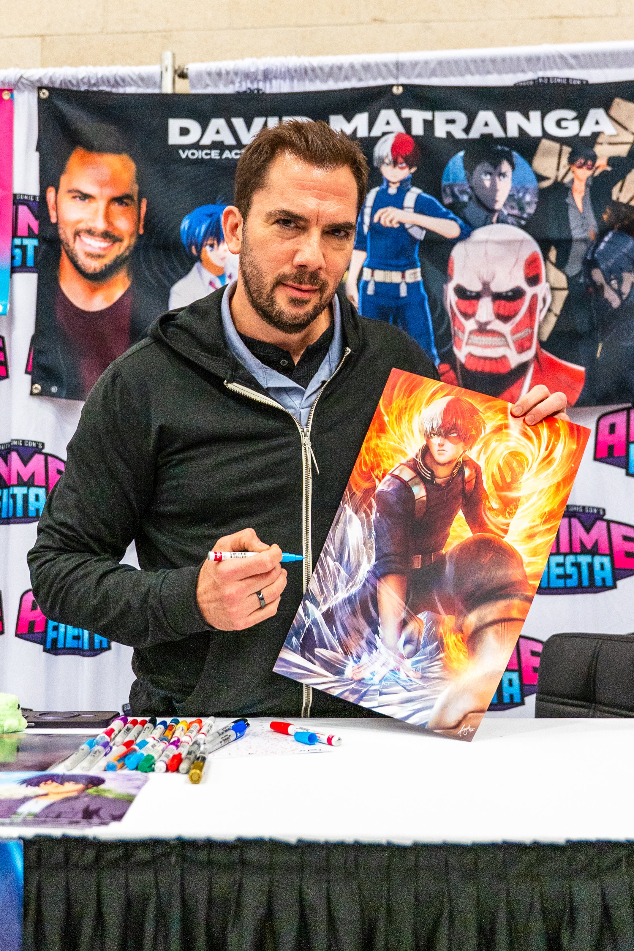 A man with brown hair and beard holding a poster with an animated character from a video game, standing in front of a colorful banner at a convention.