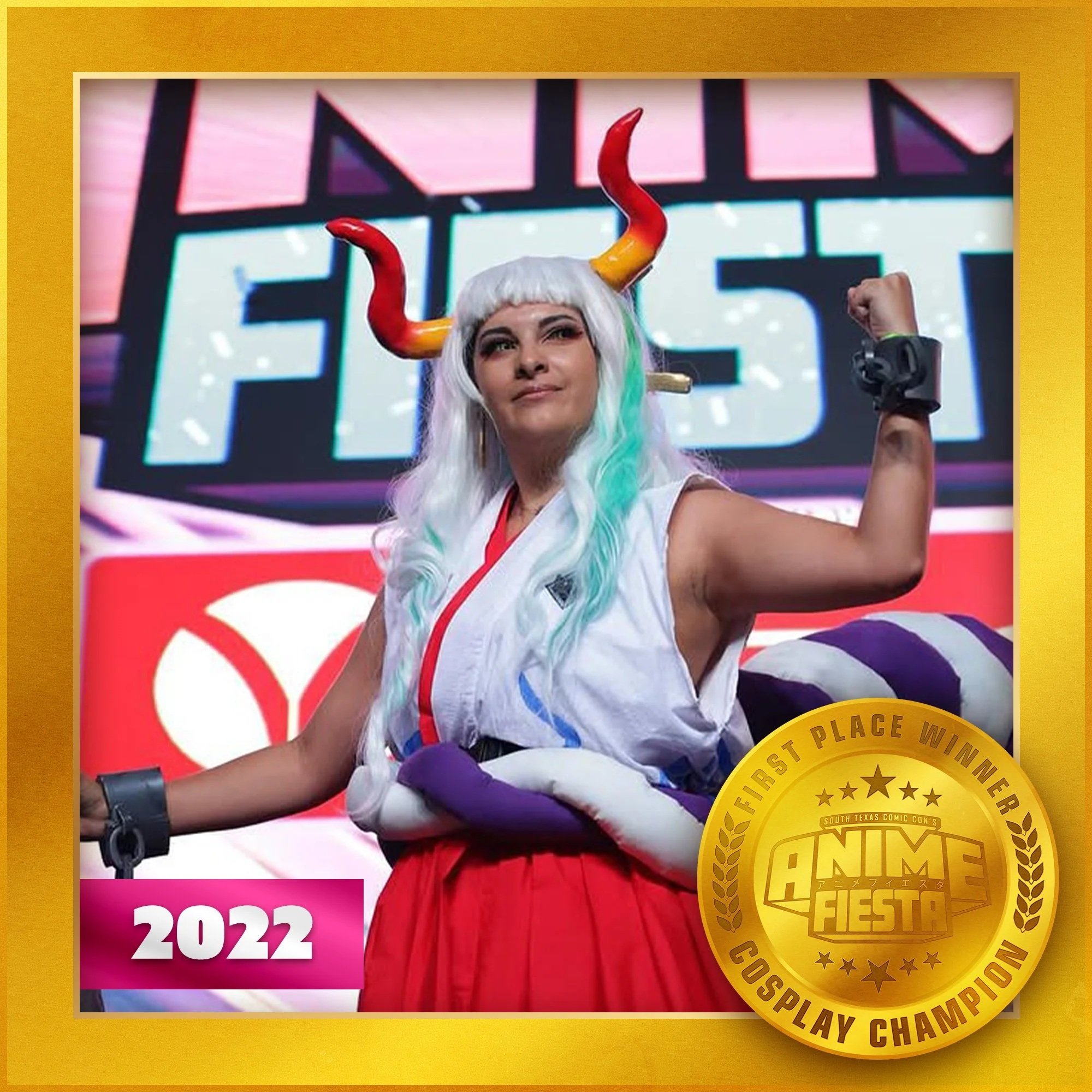 A woman dressed as a demon with white hair, red and yellow horns, and a white and red outfit, raising her fist in victory at an event with a large sign in the background and a gold award medal on the right side.