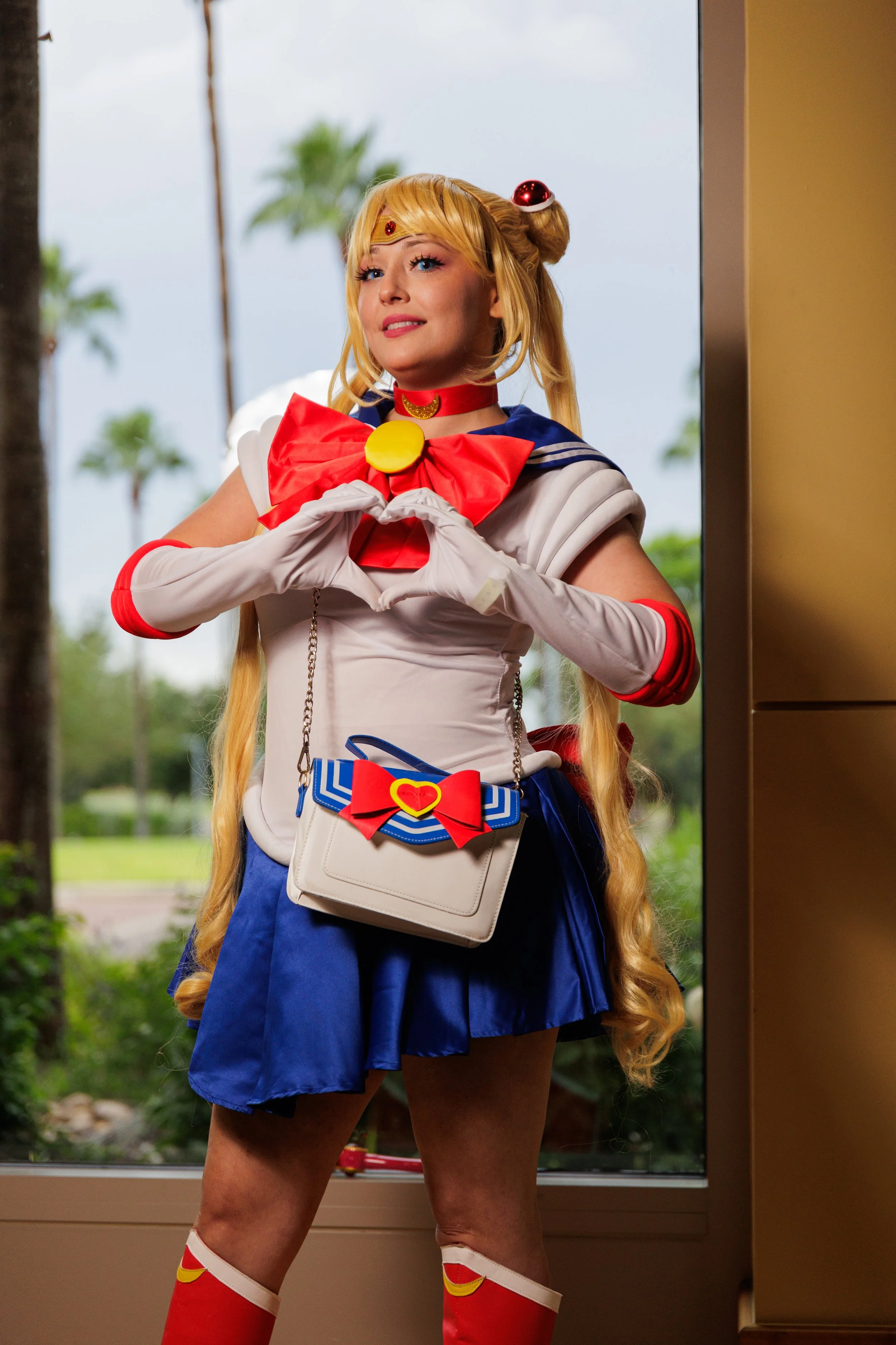 Woman dressed as Sailor Moon making a heart shape with her hands in front of a glass window with palm trees outside.