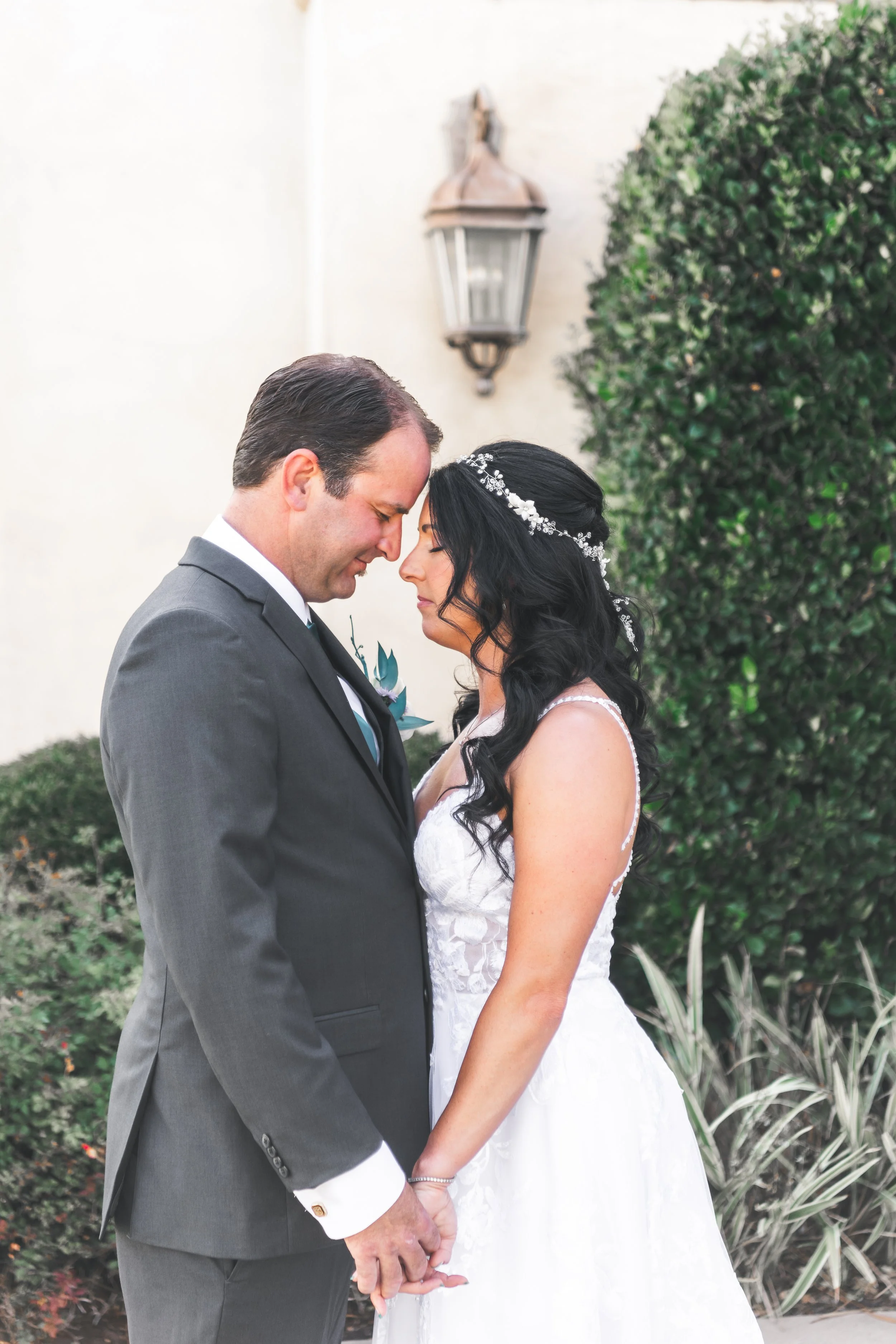 Bride and groom standing close, holding hands, with their foreheads touching in an outdoor setting.