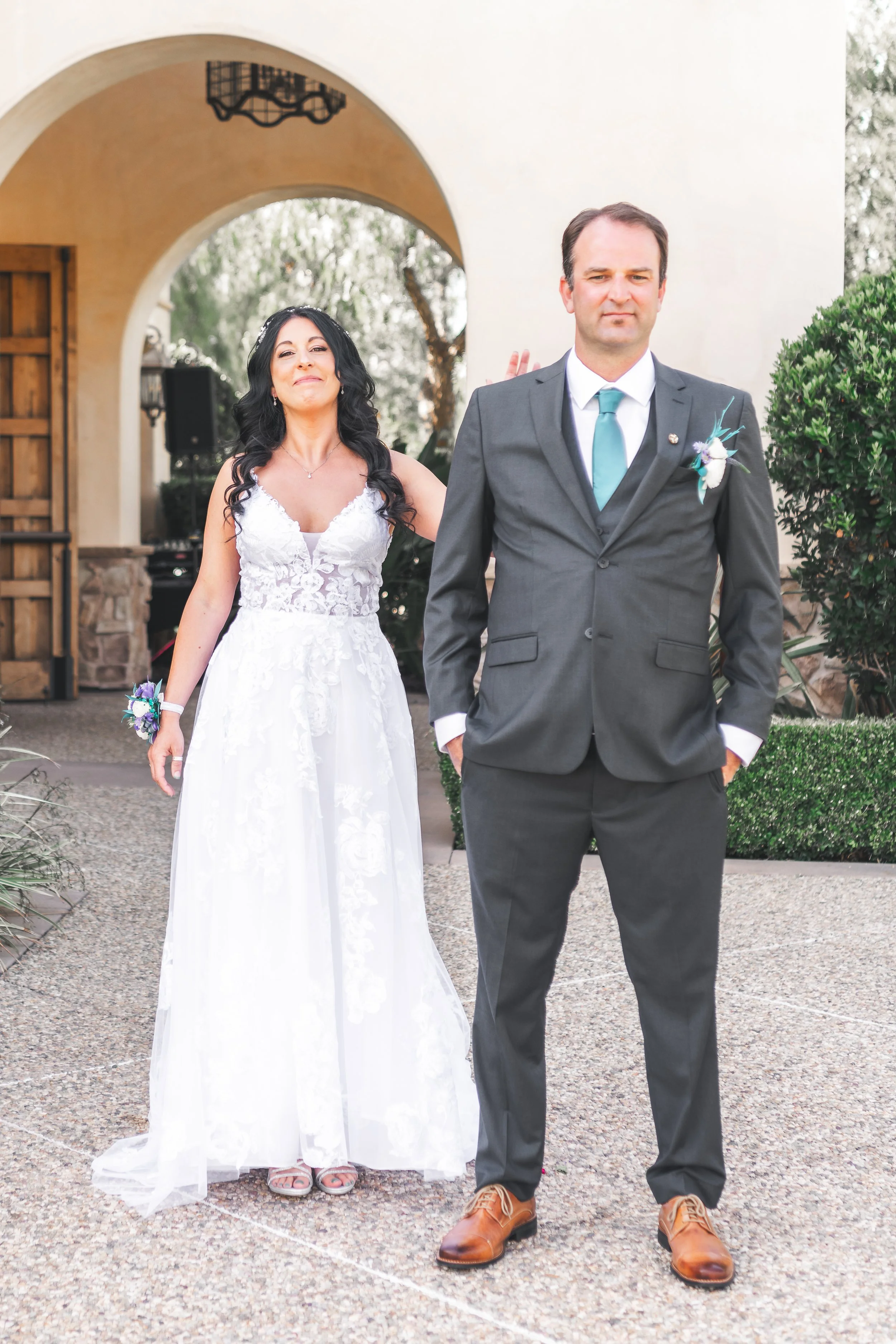 Bride in a lace wedding dress and groom in a gray suit with a teal tie standing outdoors.