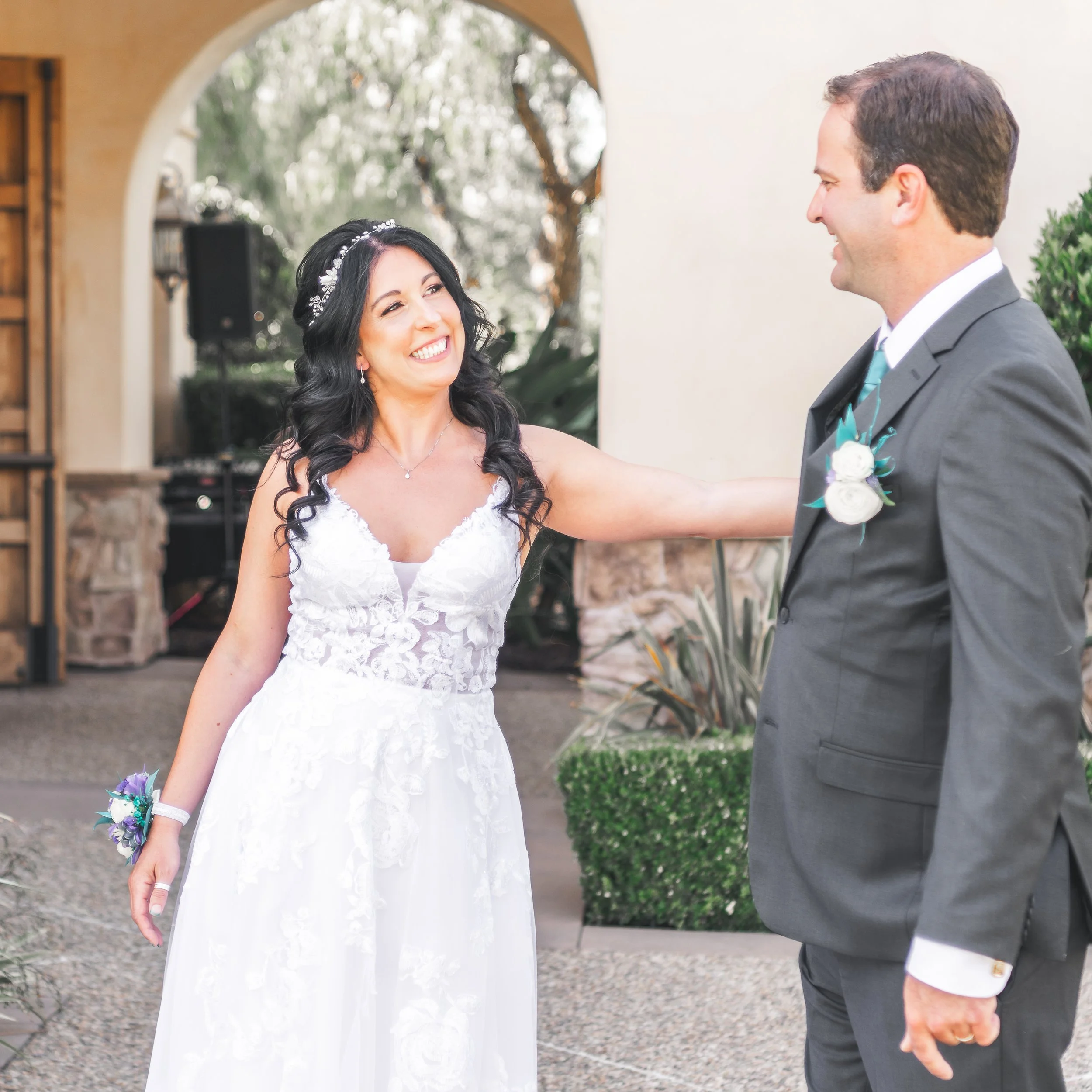 Bride in white lace wedding dress smiling at groom in dark suit, outside venue.